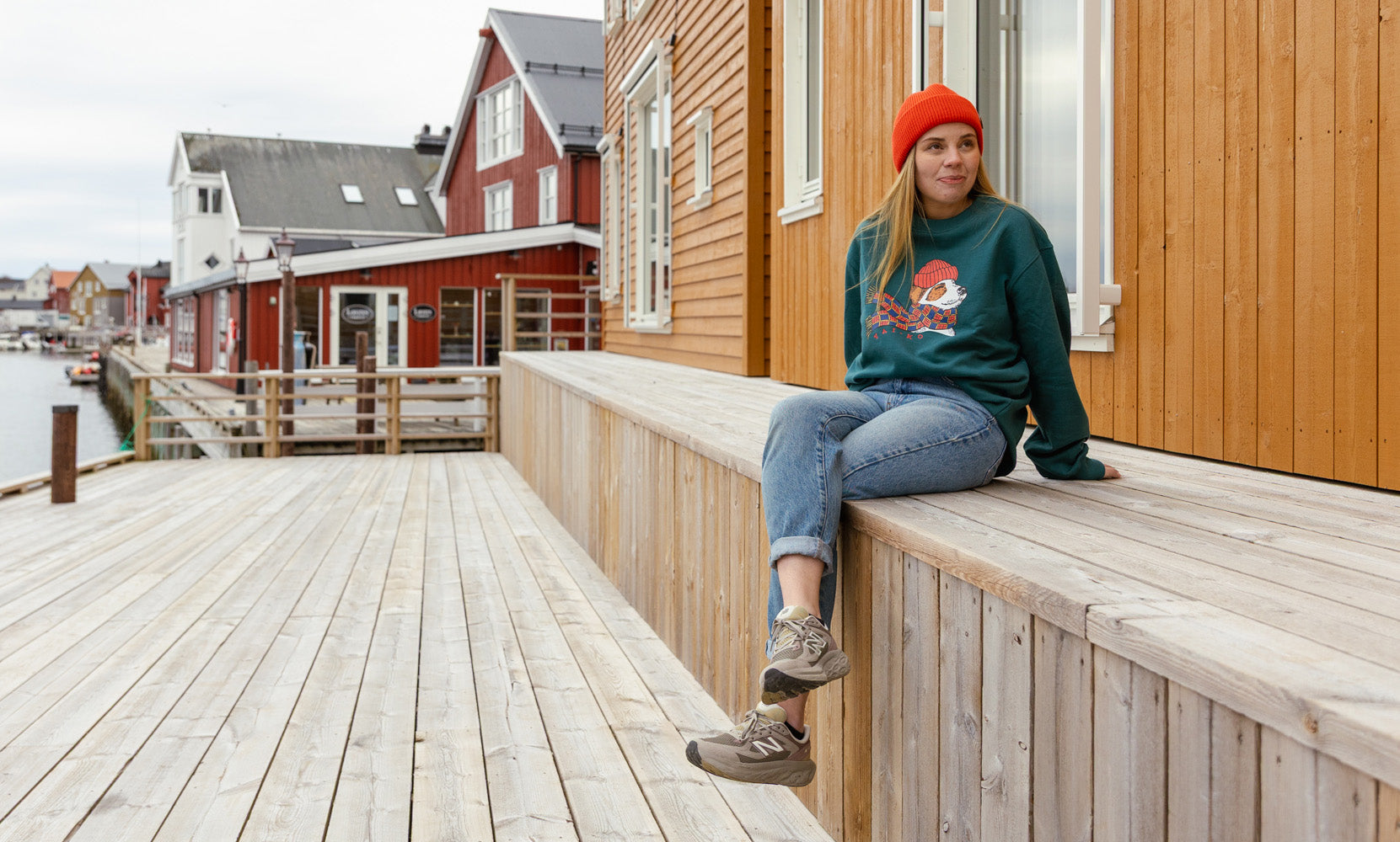 A woman in a red beanie, green sweatshirt, jeans, and sneakers sits on a wooden deck by orange and red buildings, looking relaxed. The scene appears calm and outdoors.