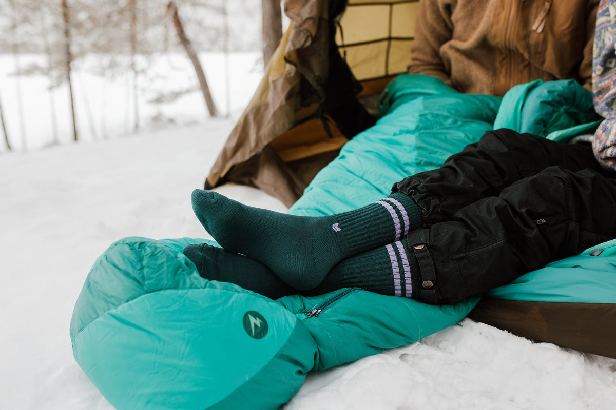 Two people relax in a teal sleeping bag inside a tent, wearing warm socks, surrounded by snow.