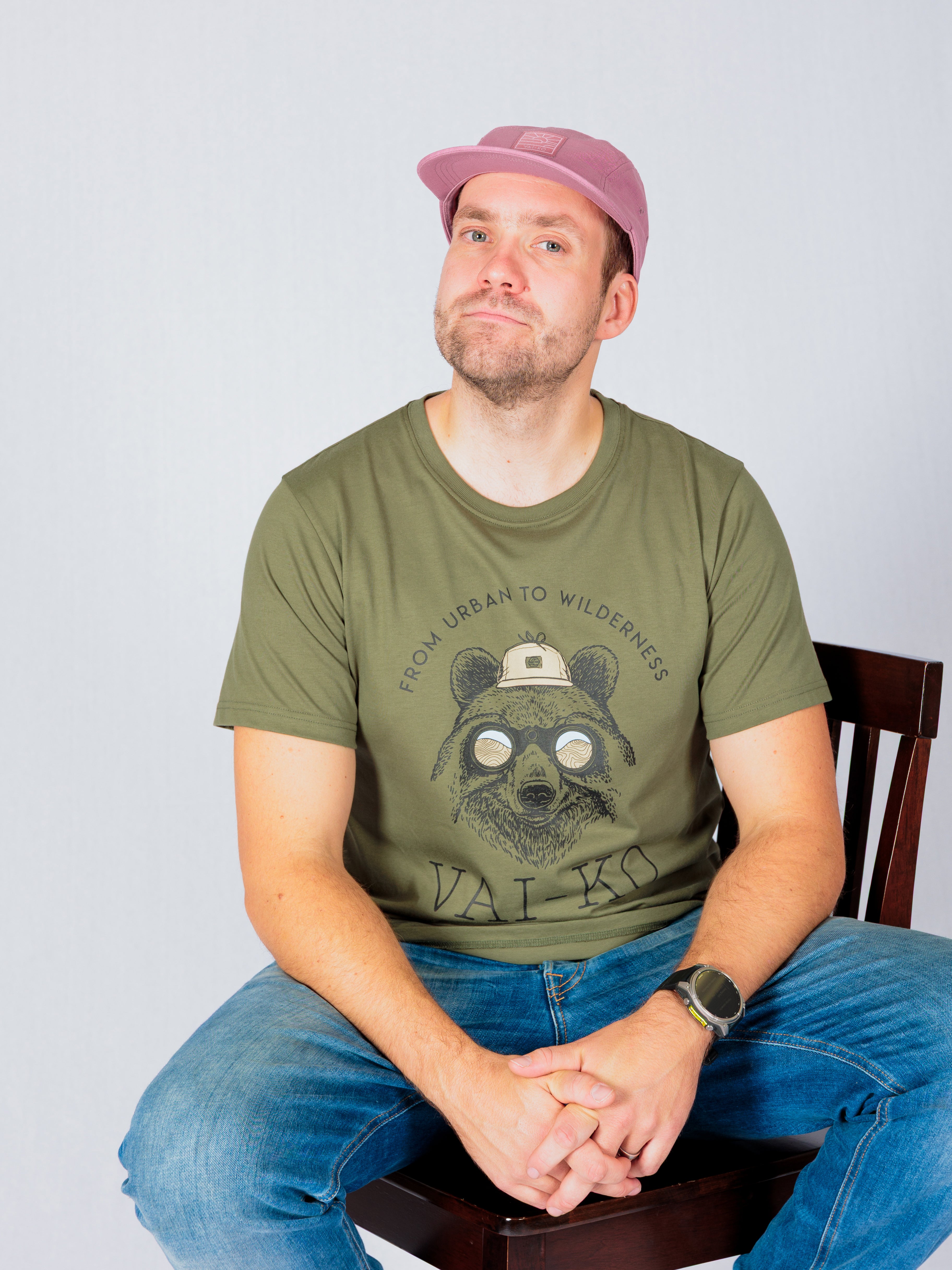 A man in a pink cap and green t-shirt with a bear graphic sits on a chair against a plain background.