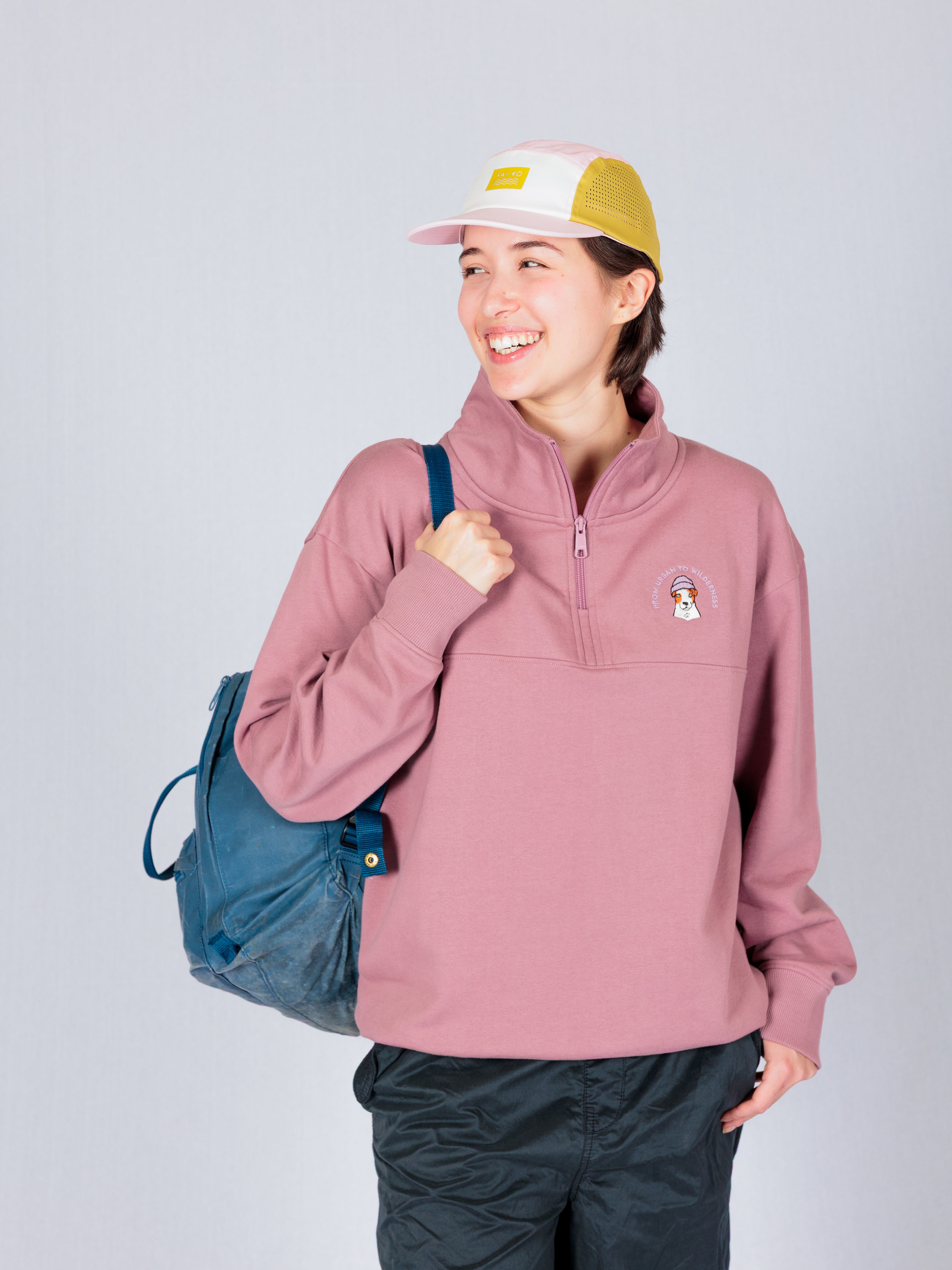 A smiling person in a pink sweatshirt and cap carries a blue backpack and looks to the side.