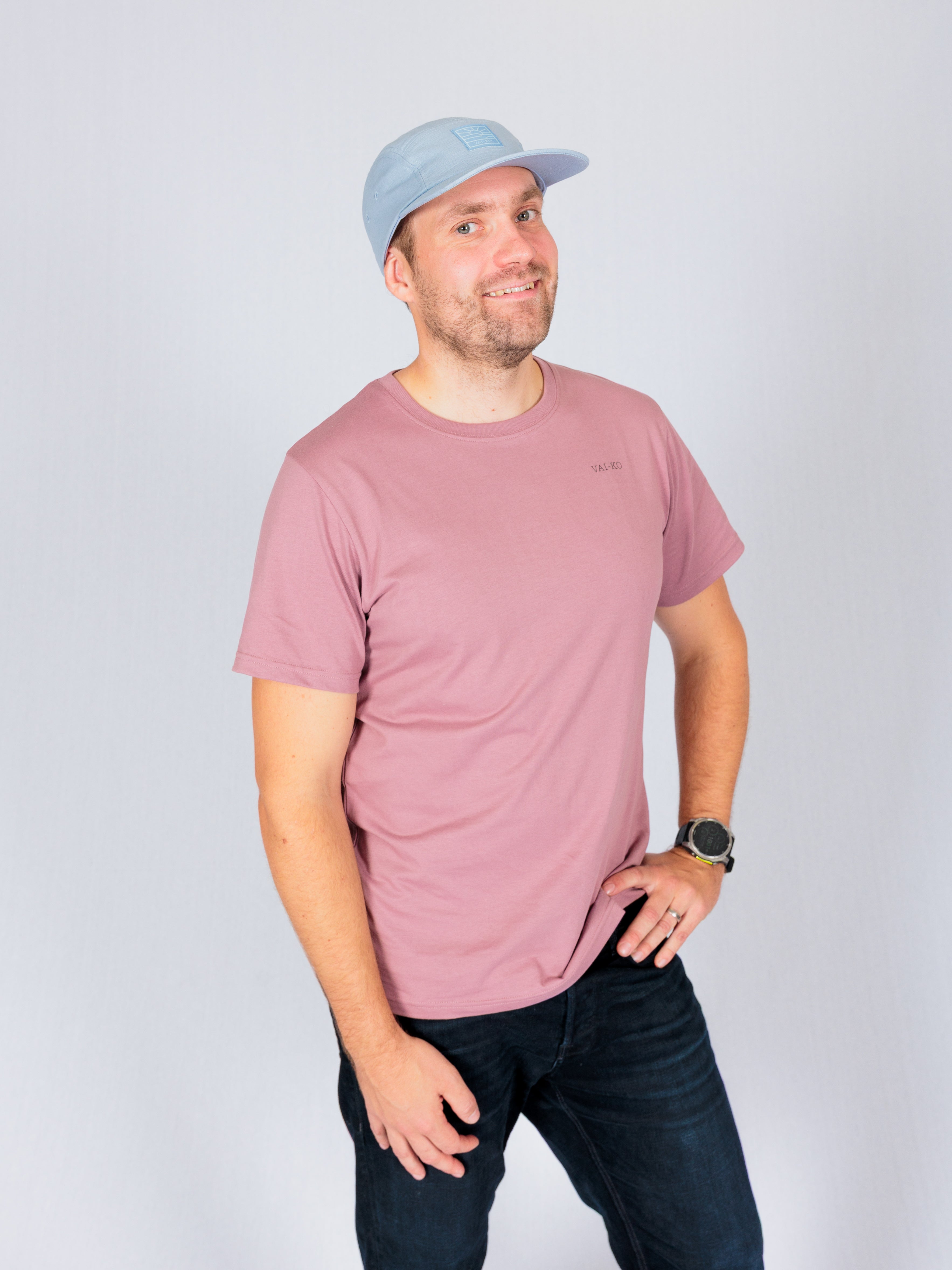 Man in a mauve t-shirt and light blue cap smiling, standing against a plain light background.