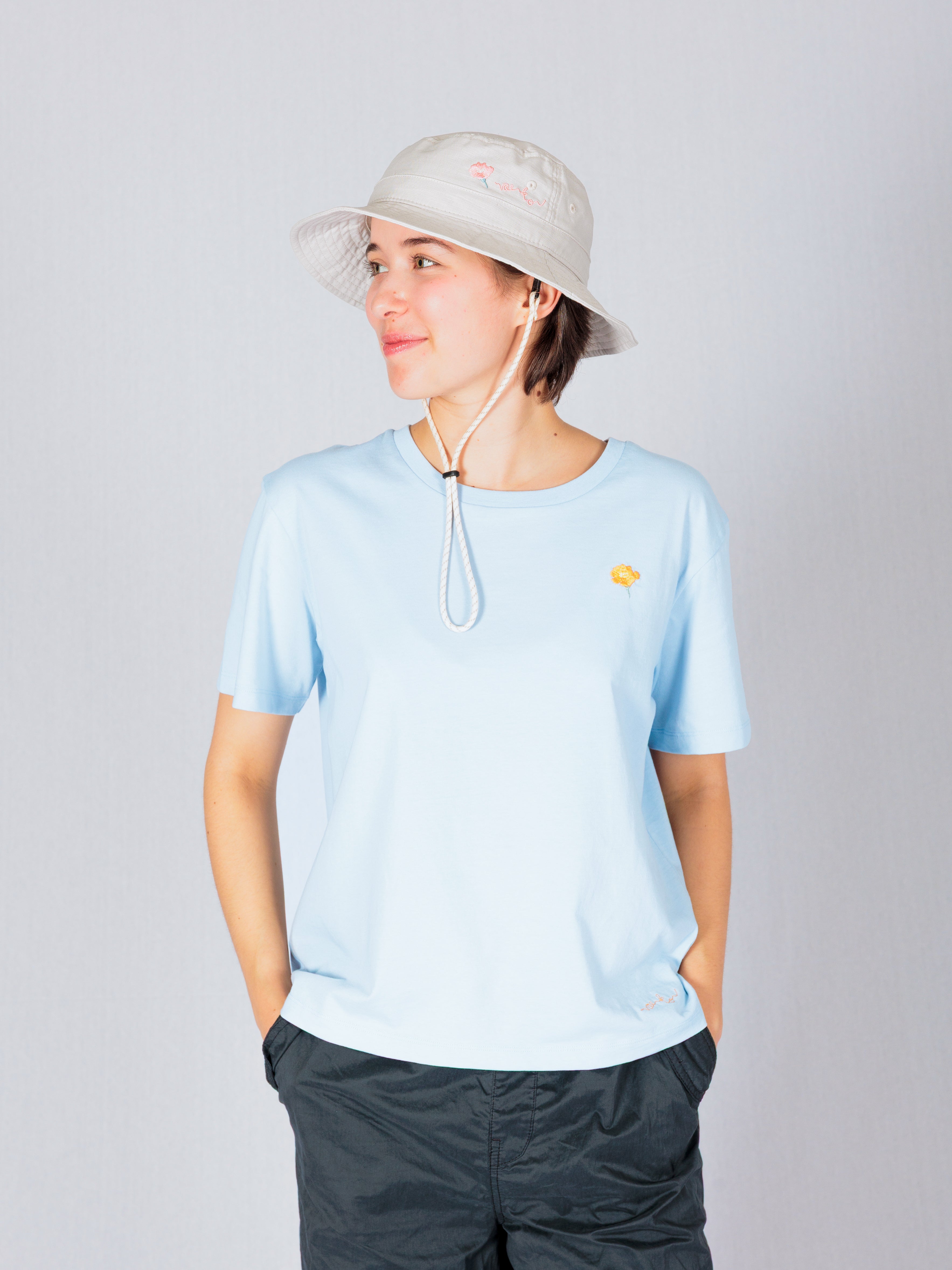 Person in a light blue shirt and bucket hat, hands in pockets, looking to the side against a plain background.