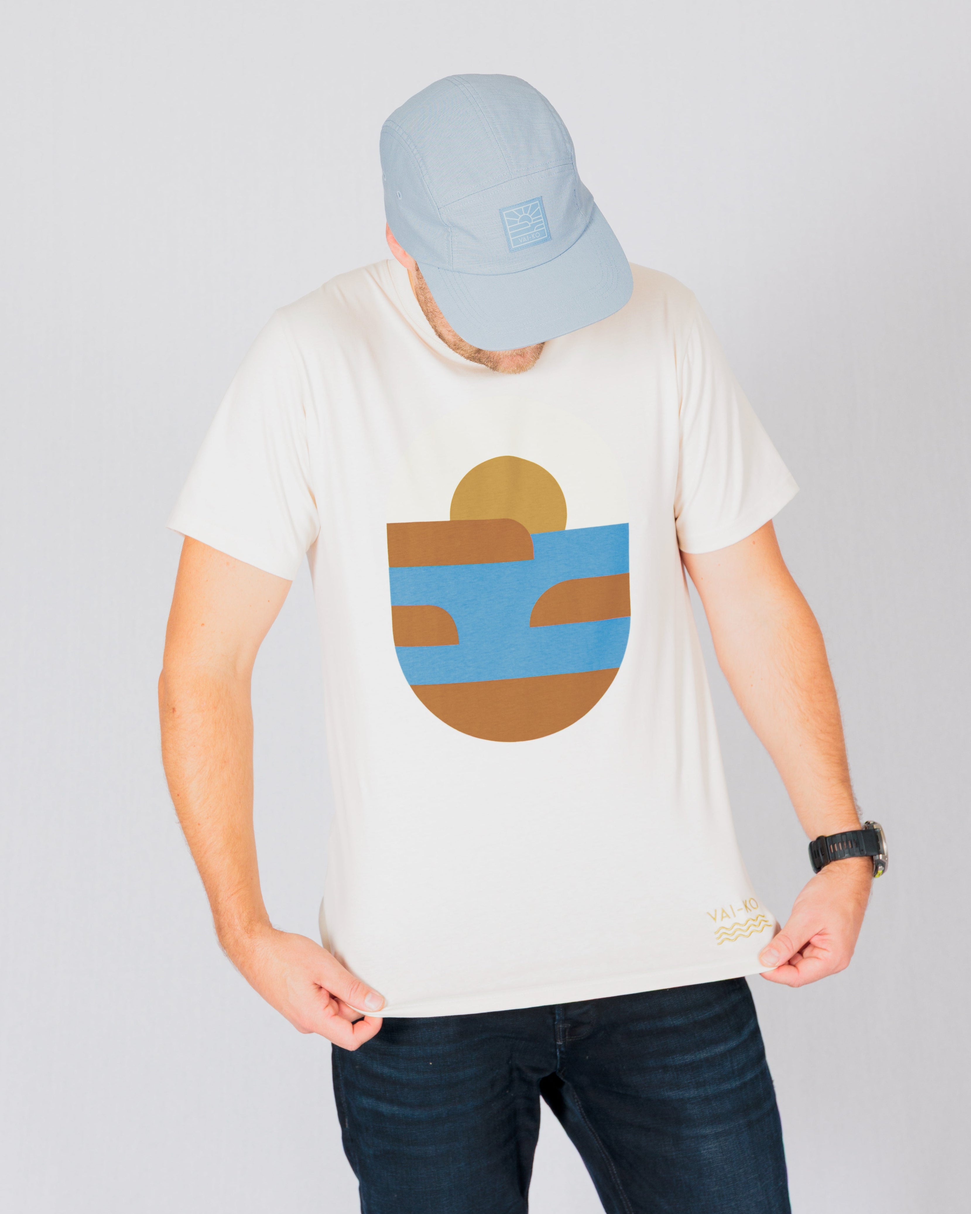 Person in a light blue cap and white t-shirt with abstract blue and brown design looks down at the shirt.