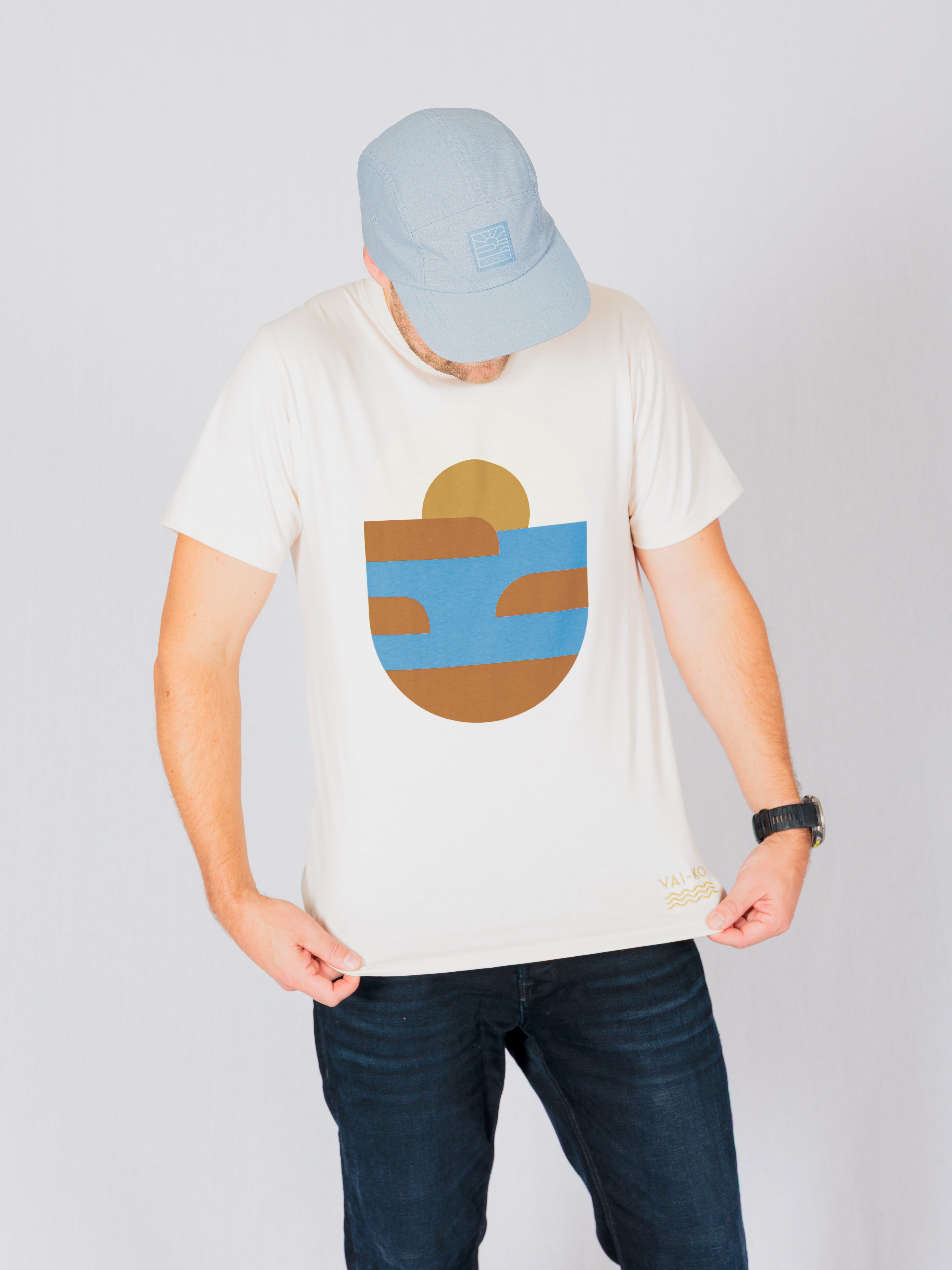Person in a light blue cap and white t-shirt with abstract blue and brown design looks down at the shirt.