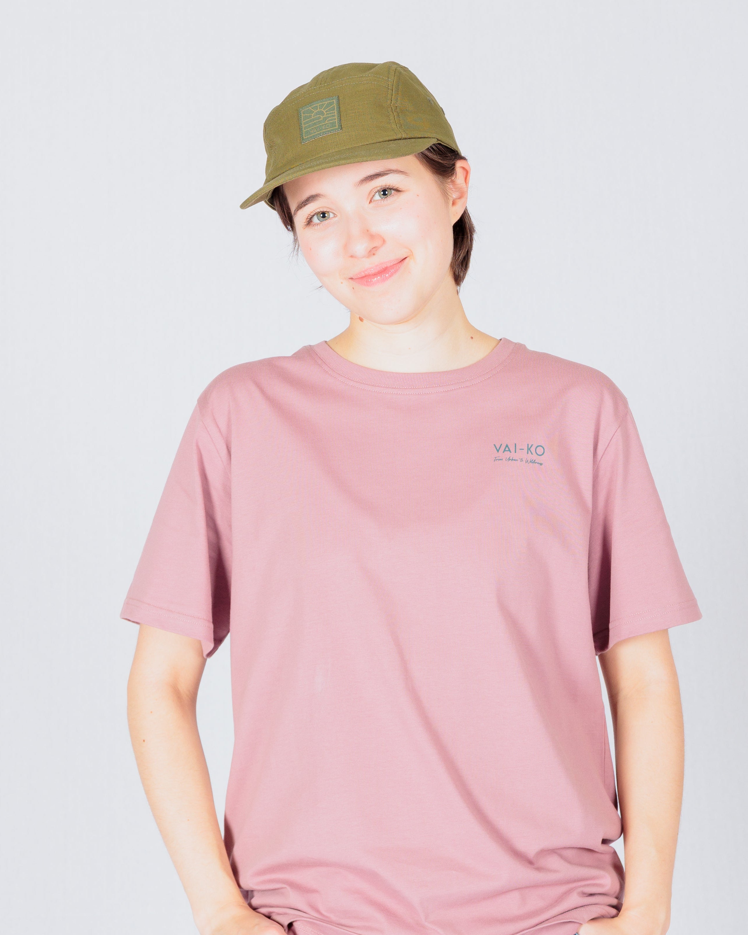 Person in a pink t-shirt, blue jeans, and olive cap smiling against a plain light background.