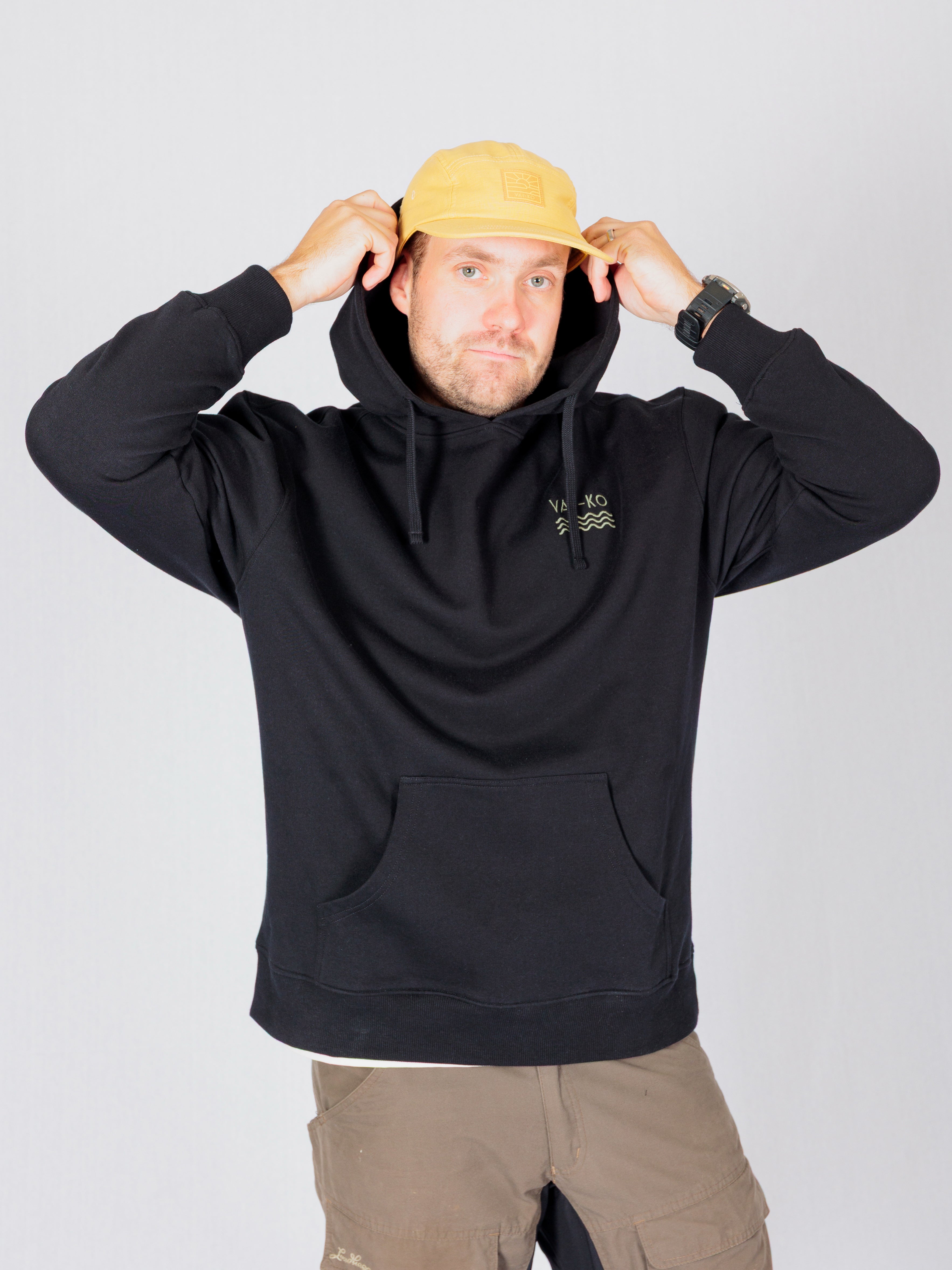 Man in a black hoodie and yellow cap adjusts his hood, standing against a plain light background.