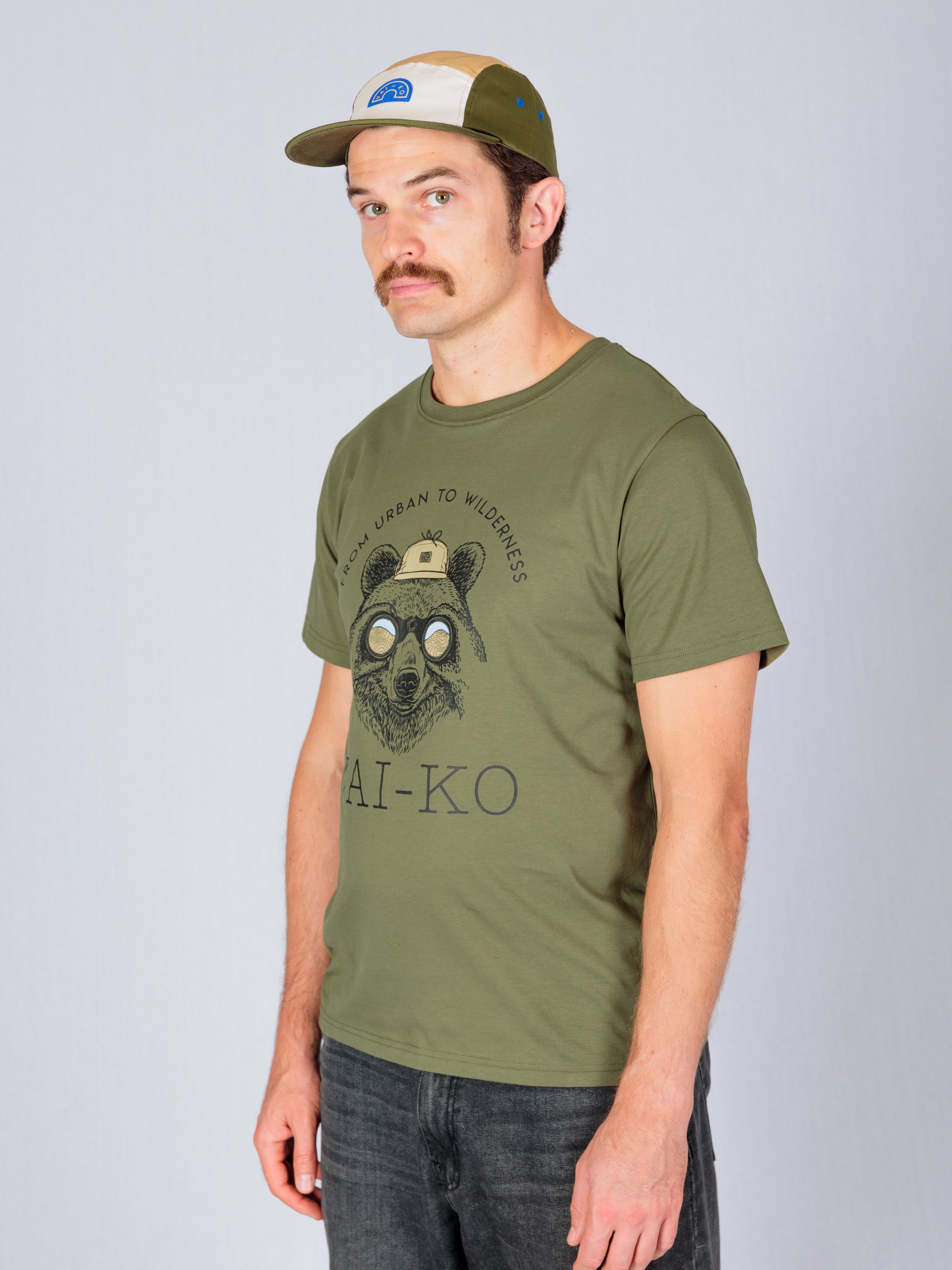 Man in an olive green t-shirt with a bear graphic and a green and beige cap stands against a plain background.