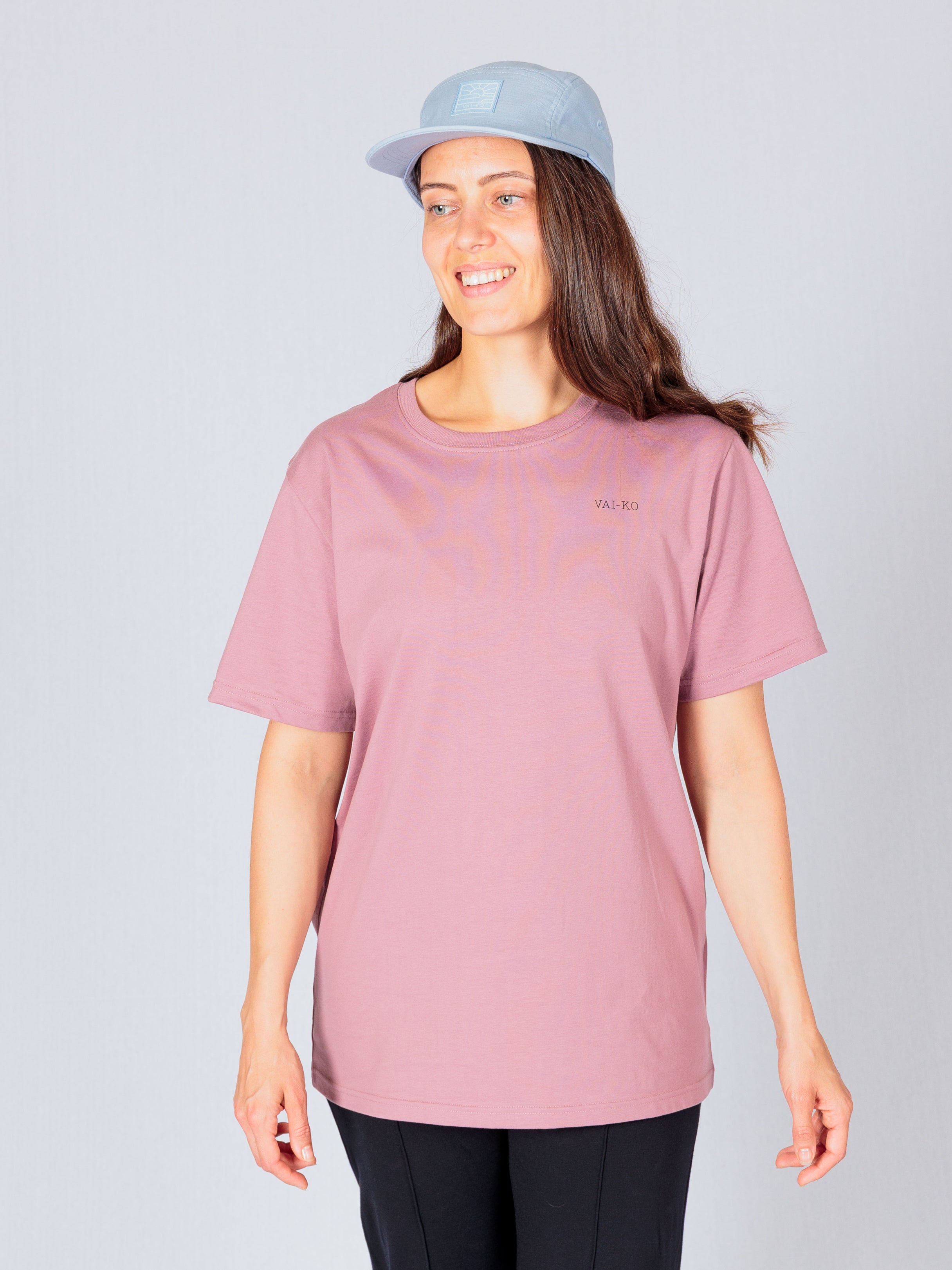 Woman wearing a mauve t-shirt, black pants, and a light blue cap, smiling against a plain background.