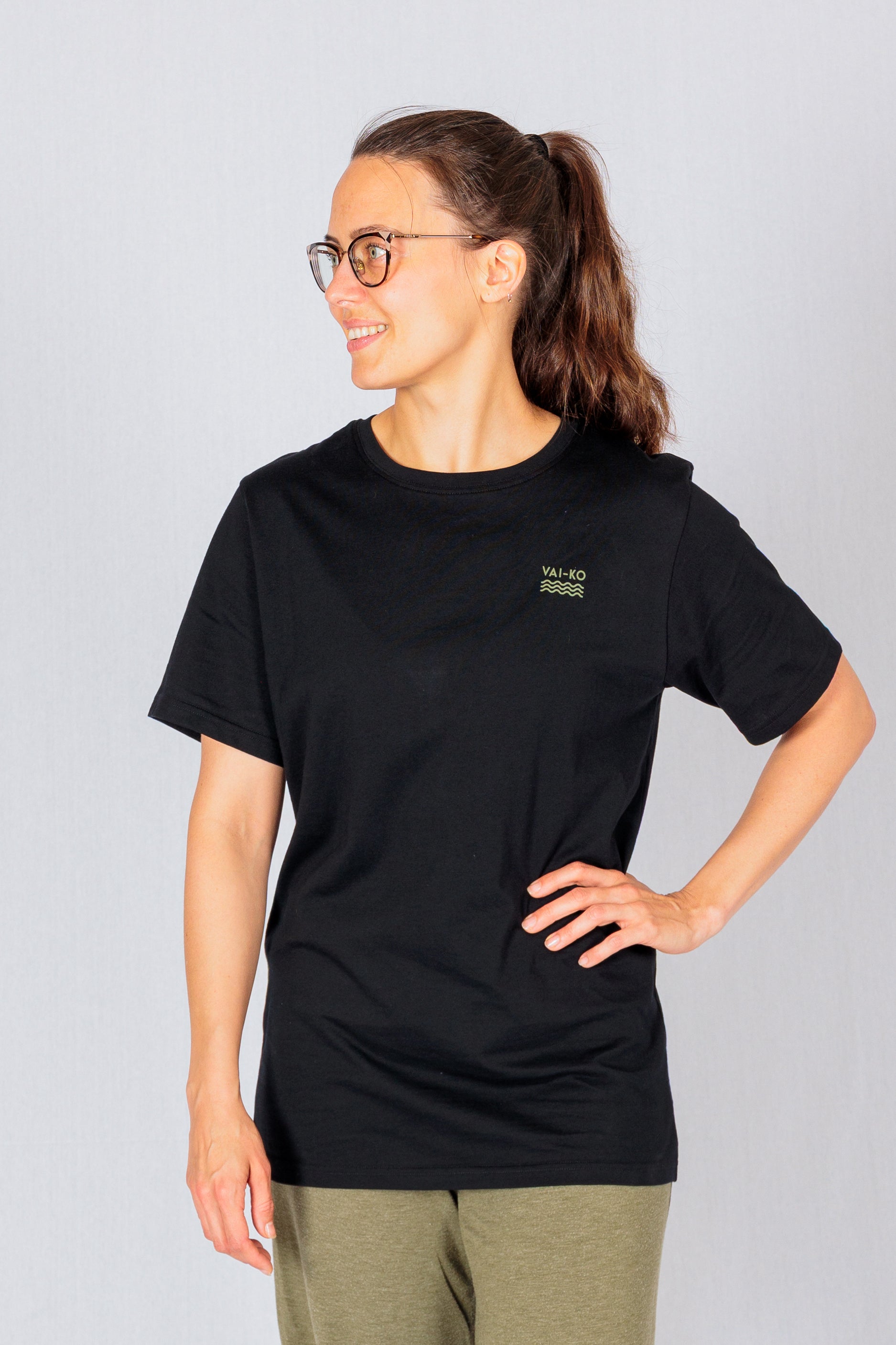Woman in glasses wearing a black T-shirt and green pants stands smiling with one hand on her hip.