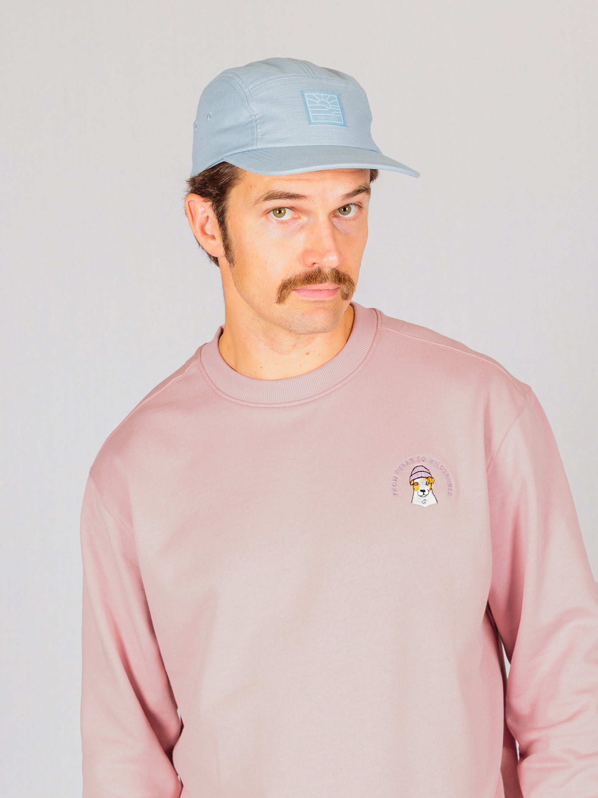 Man with mustache wearing a light blue cap and pink sweatshirt against a light background.