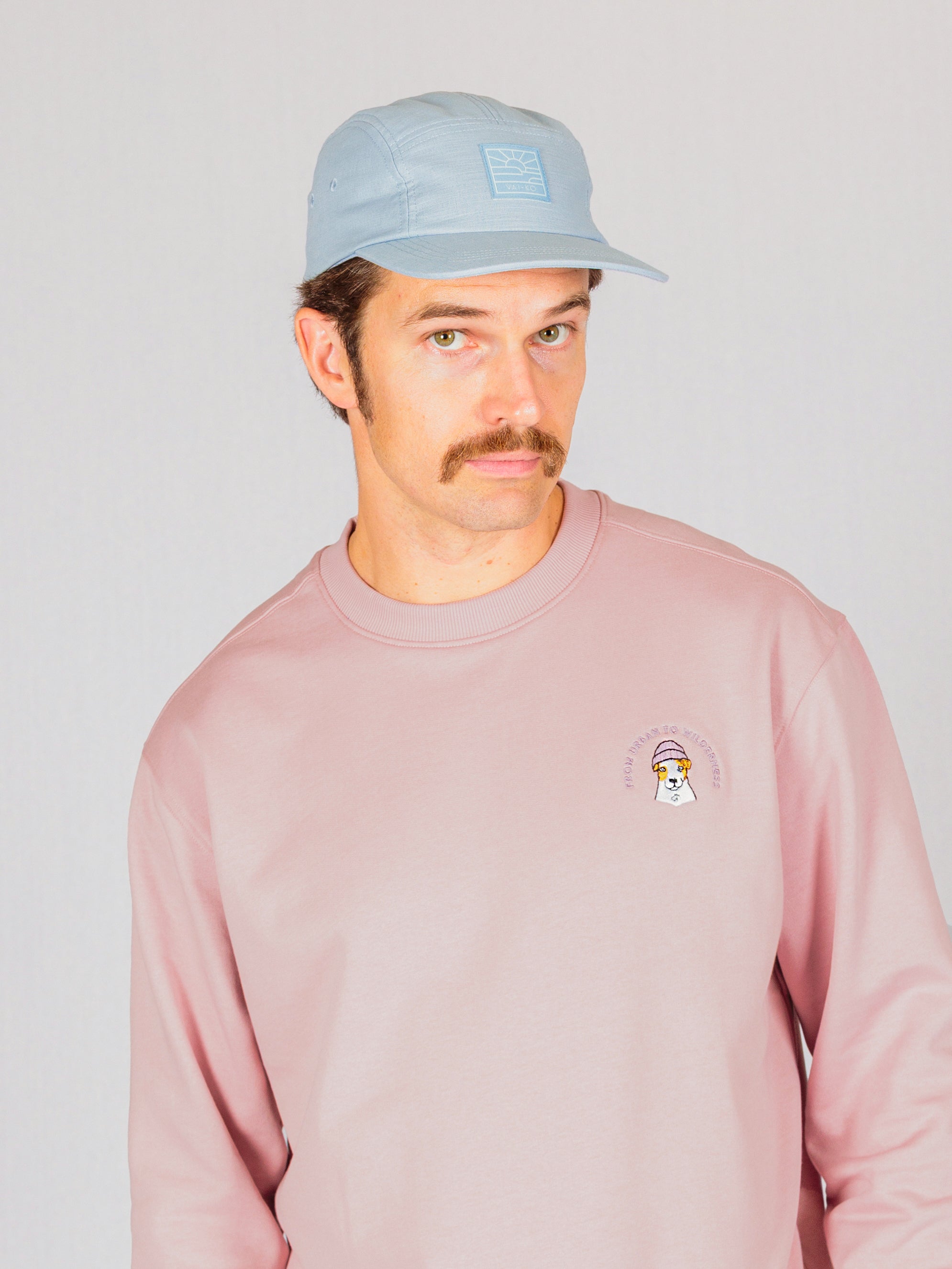 Man with mustache wearing a light blue cap and pink sweatshirt against a light background.