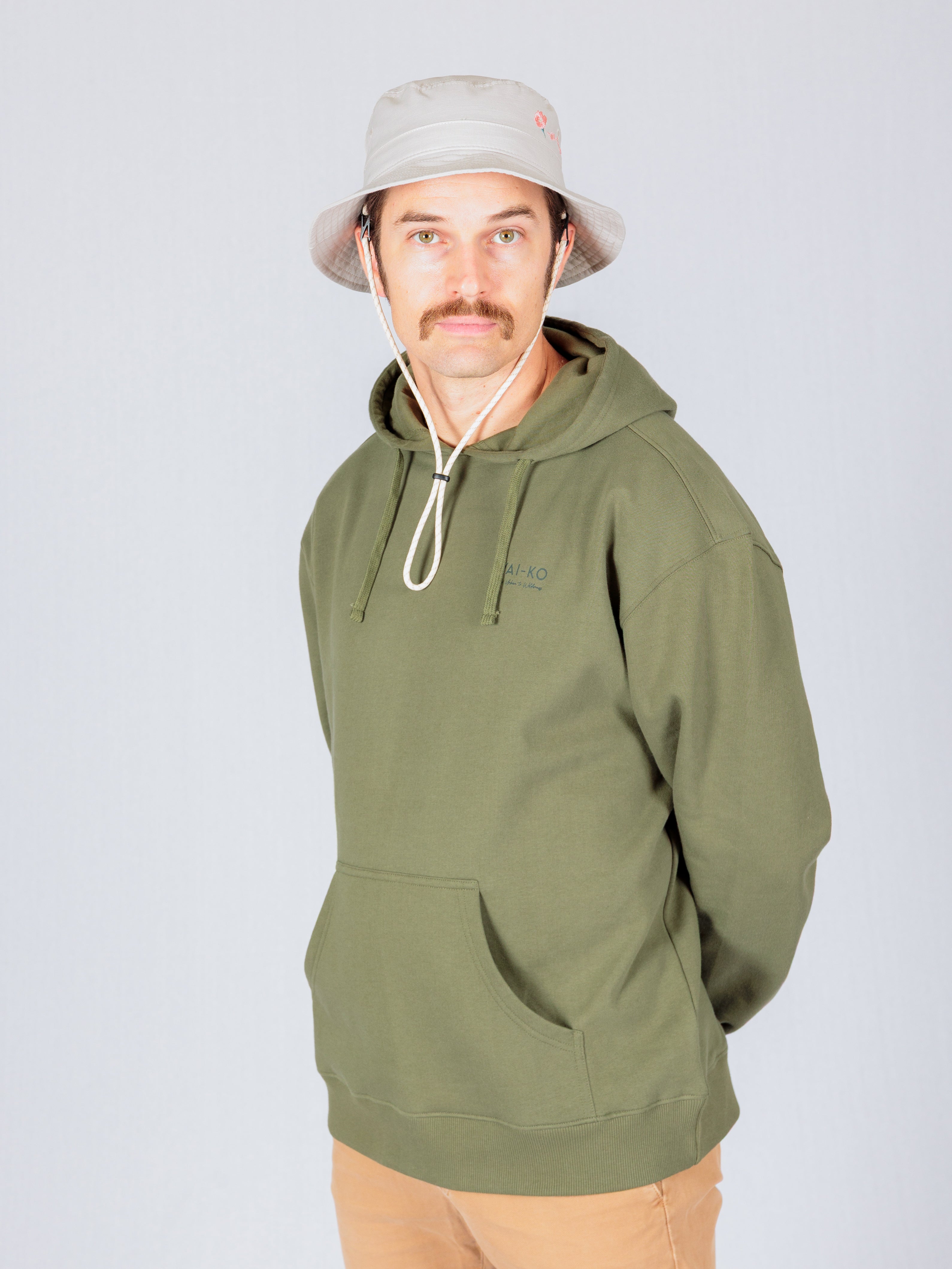 Man in olive green hoodie and beige bucket hat standing against a plain light background, looking at the camera.