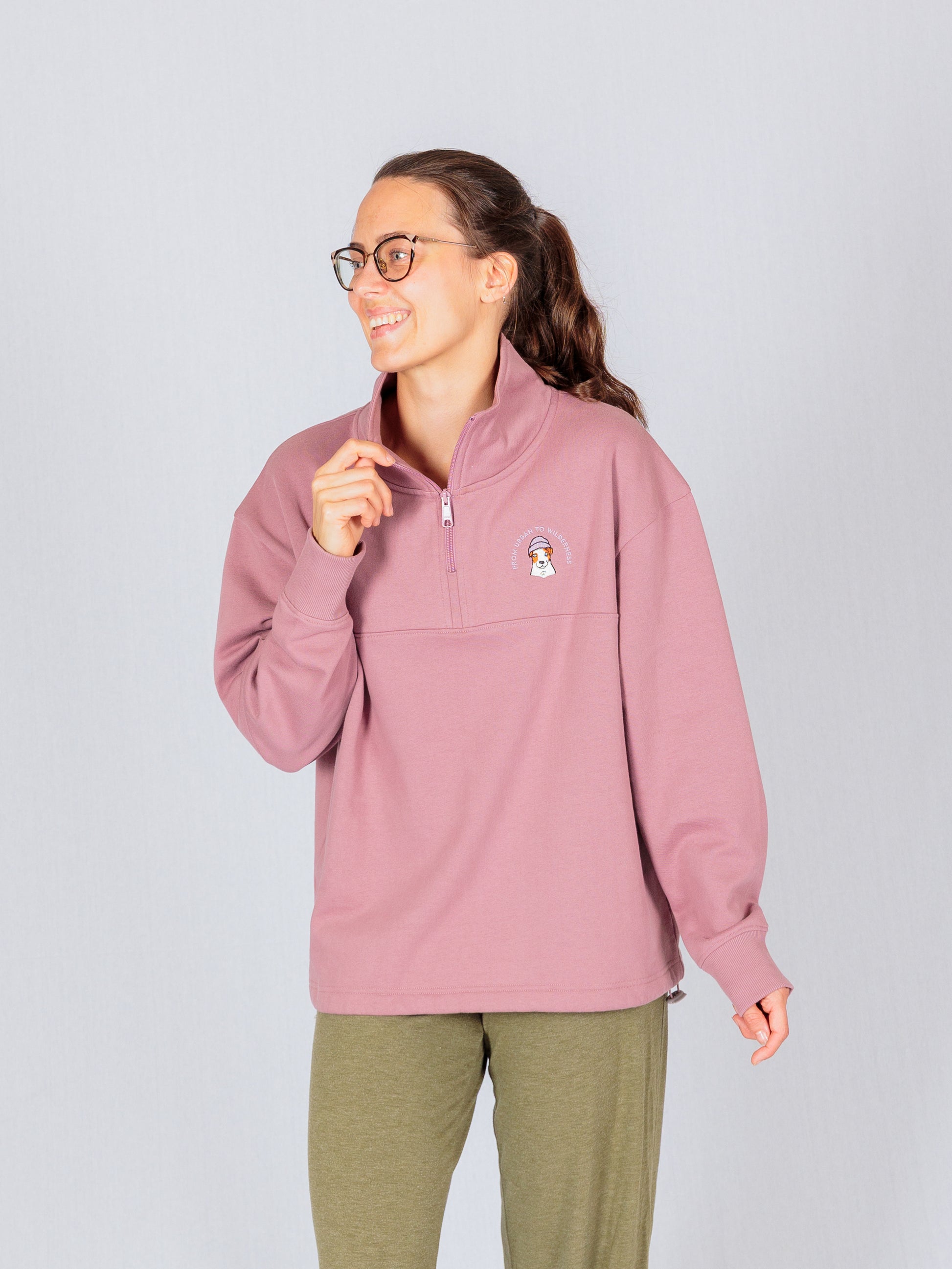 Smiling woman in glasses wearing a mauve quarter-zip sweatshirt and olive pants, standing against a light background.