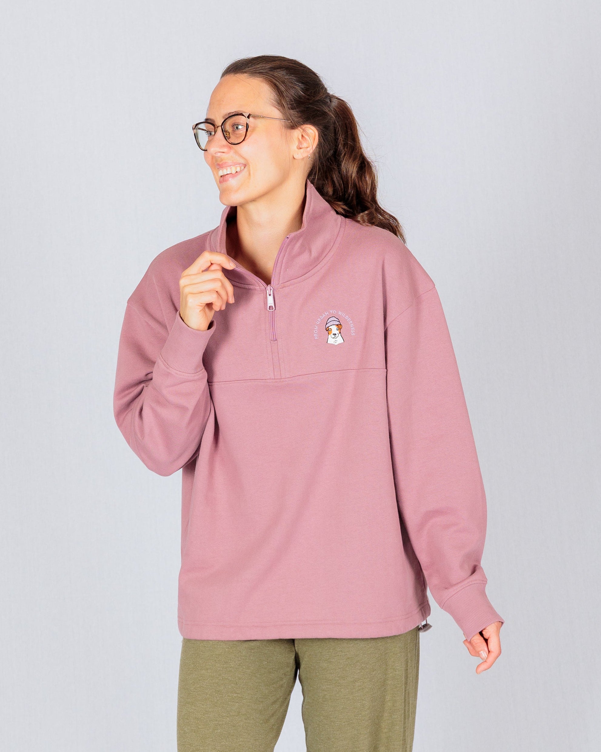 Smiling woman in glasses wearing a mauve quarter-zip sweatshirt and olive pants, standing against a light background.