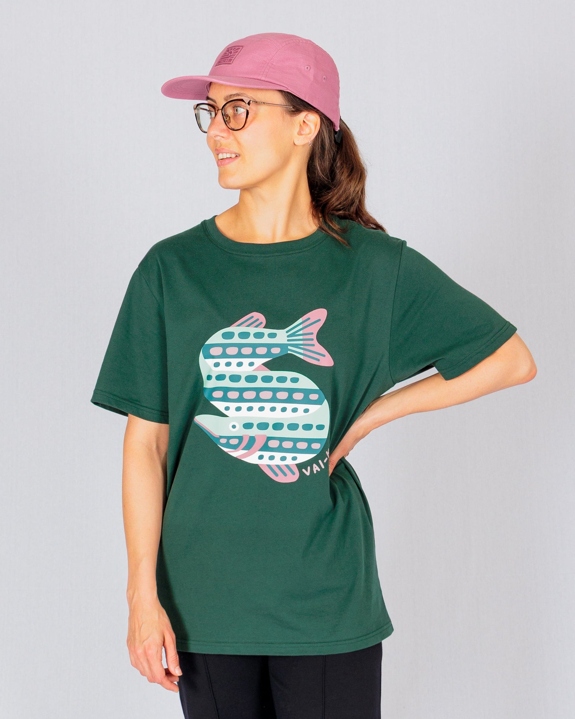 Woman in glasses and a pink cap wearing a green t-shirt with a large, stylized fish graphic, standing sideways.