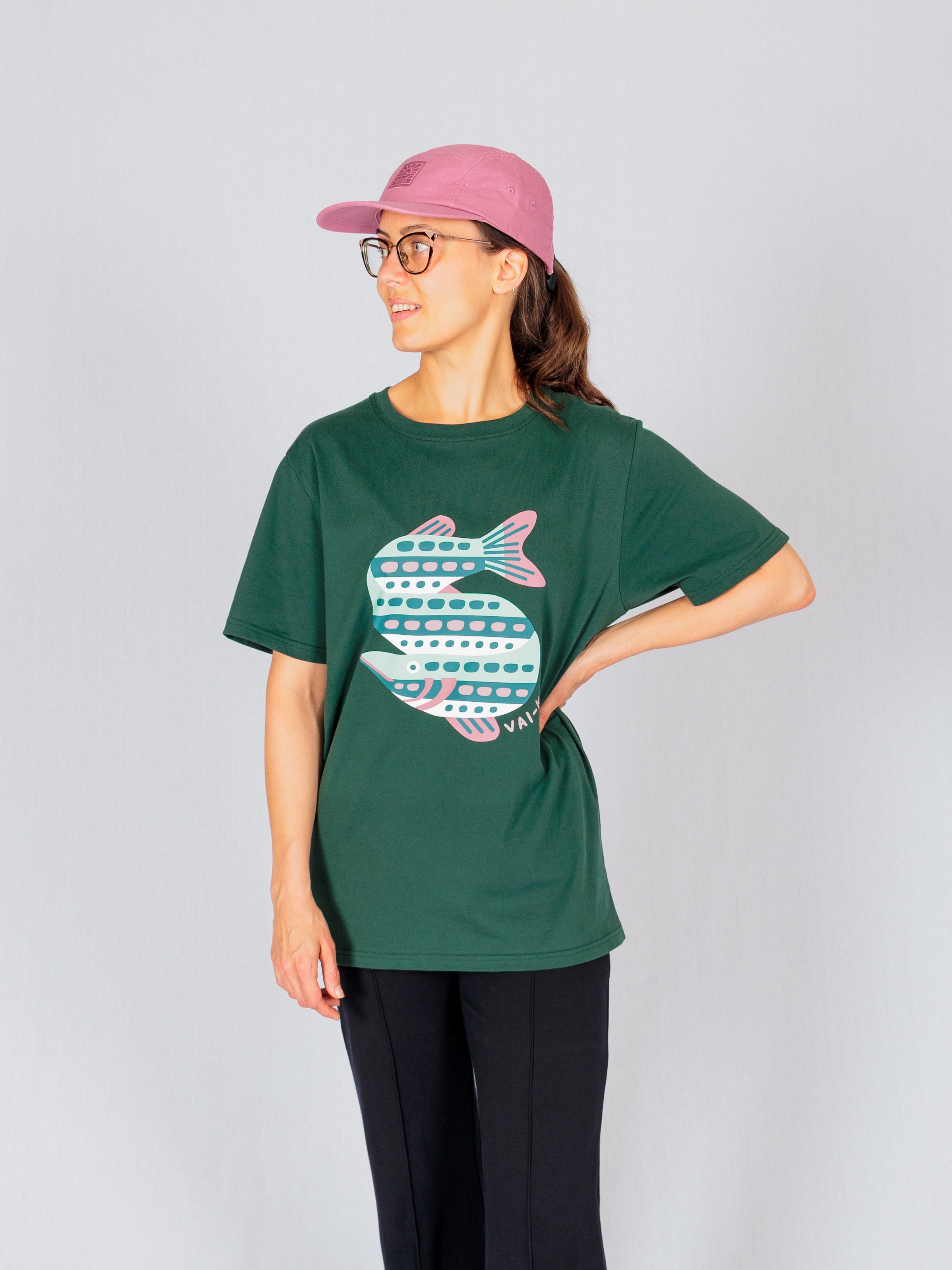 Woman in glasses and a pink cap wearing a green t-shirt with a large, stylized fish graphic, standing sideways.
