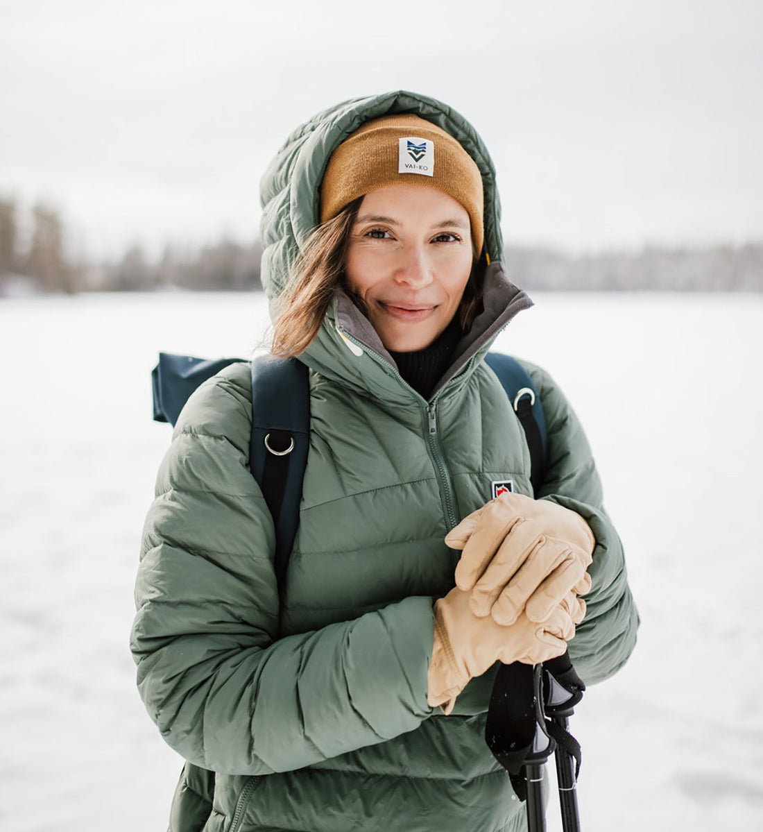 Woman in a green winter jacket and brown beanie standing outside in snowy landscape, holding ski poles.