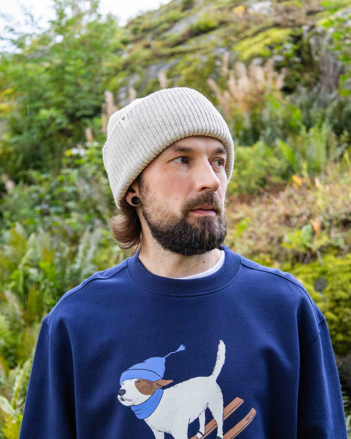 Man with a beard wearing a knit hat and blue sweater with a skiing dog graphic, standing outdoors.