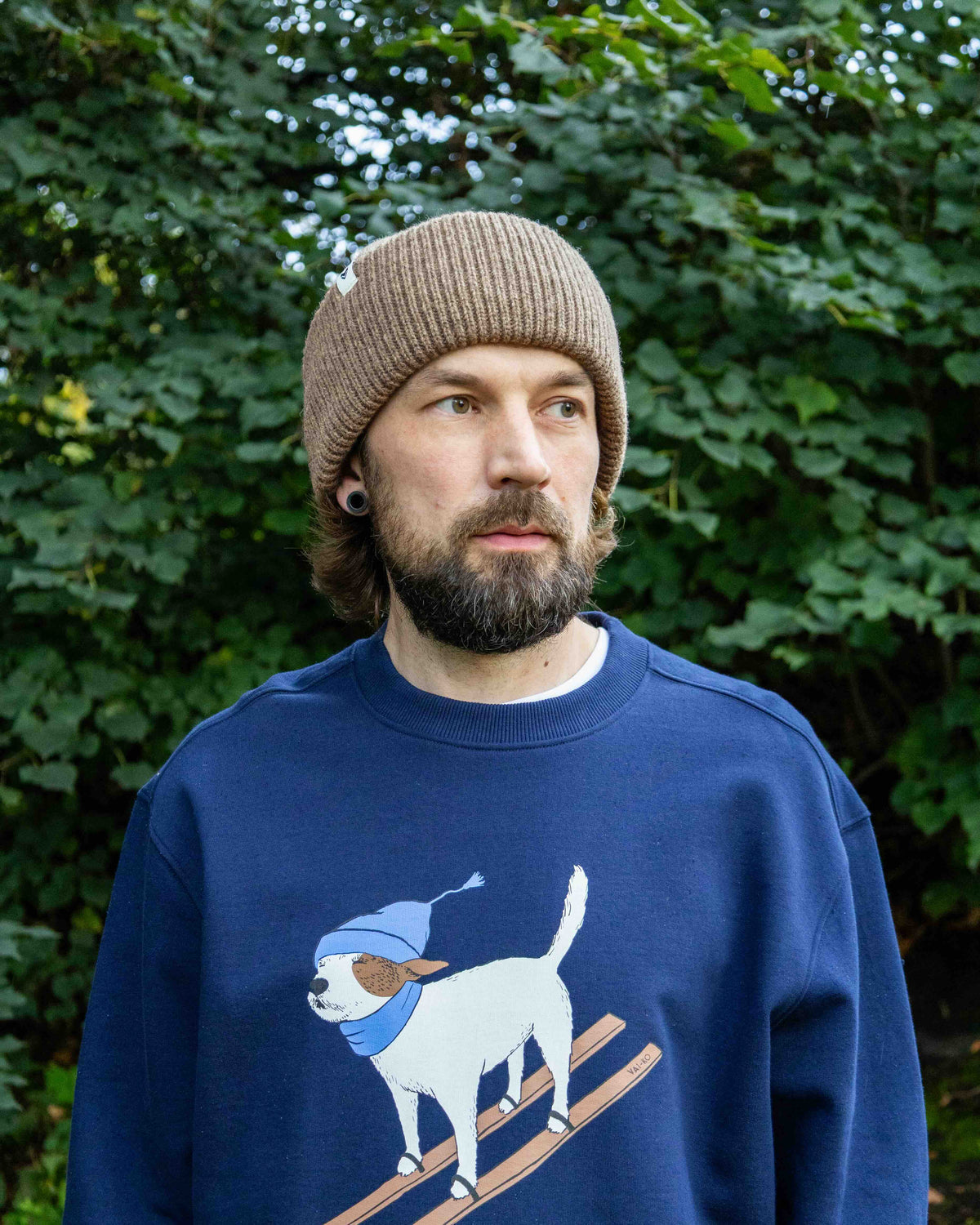 Man in a brown knit beanie and blue sweater with cartoon dog, standing in front of green foliage.