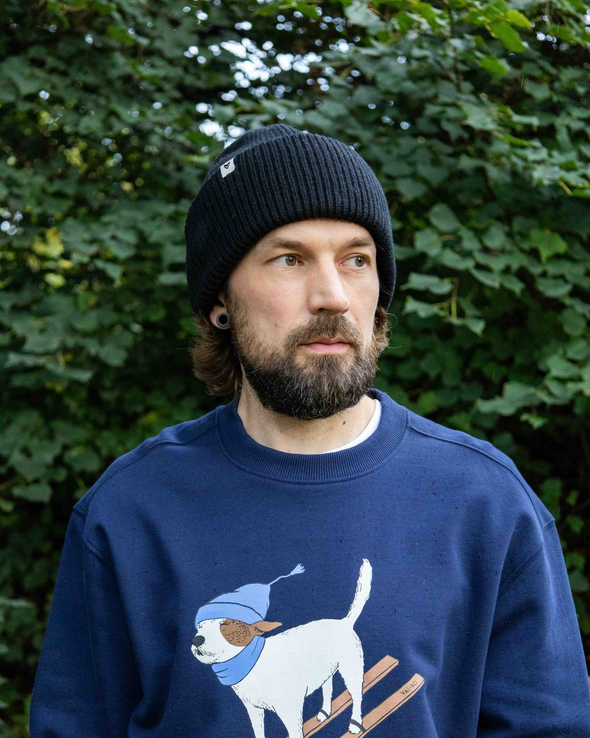 Man with a beard in a black knit hat and blue sweatshirt stands outdoors in front of green leaves.