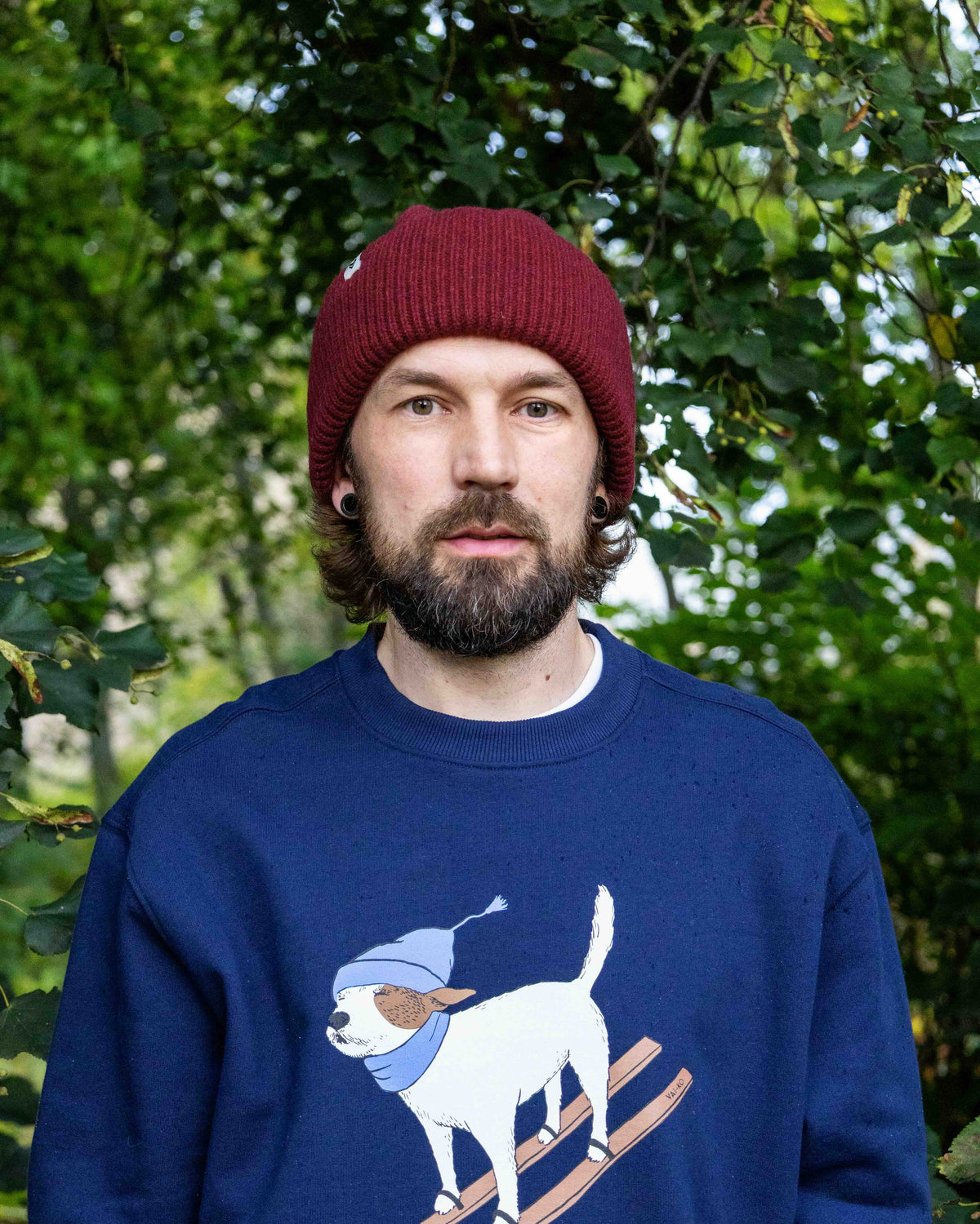 Bearded person in a red beanie and blue sweater with a skiing dog print, standing in leafy outdoor area.