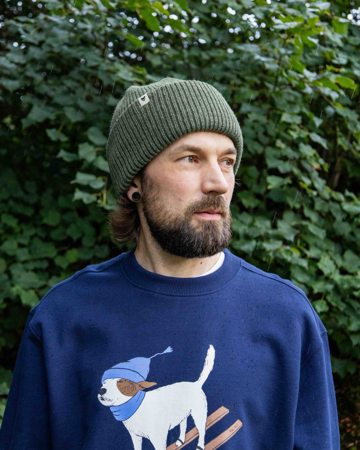 Man with a beard wearing a green beanie and blue sweater with dog graphic, standing by leafy bushes.