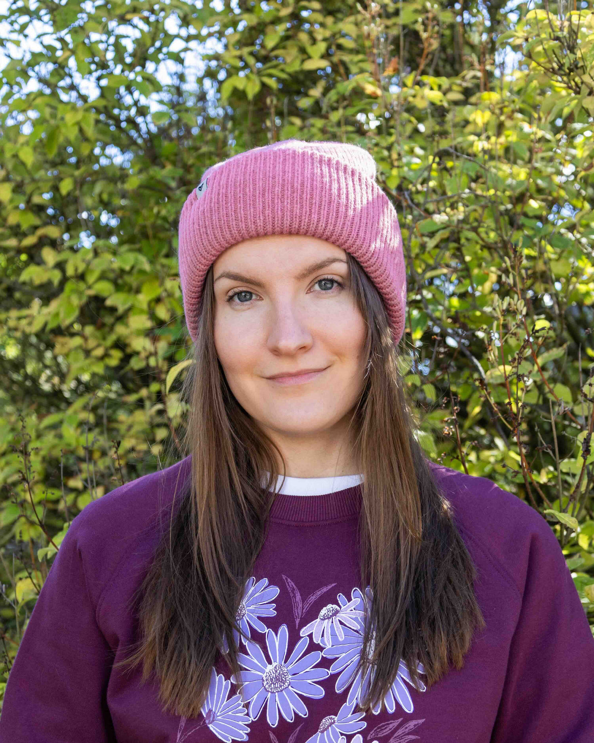 Woman with long brown hair in a pink beanie and purple sweater stands outside in front of green foliage.