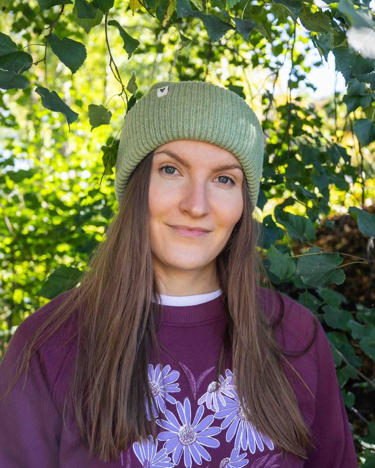 Woman with long brown hair, green beanie, and maroon sweater stands outside among green leaves and sunlight.