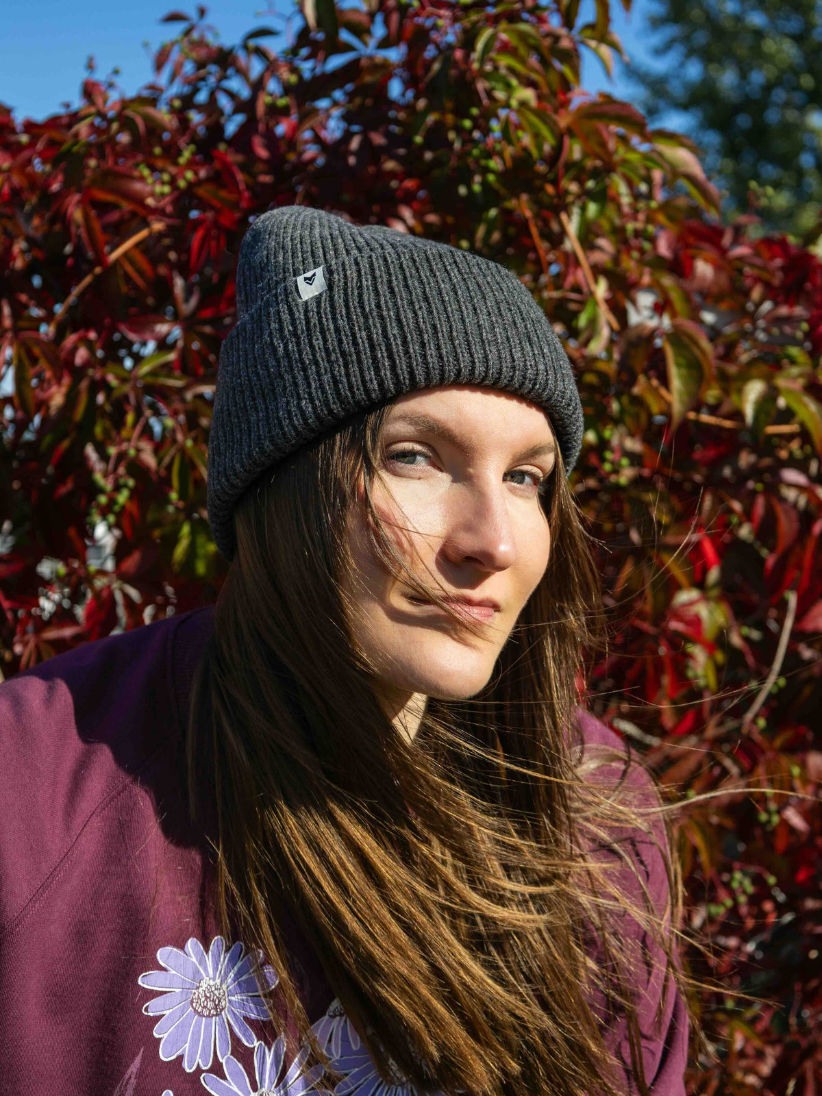 Woman in a gray knit beanie and maroon top stands outdoors in front of red foliage on a sunny day.