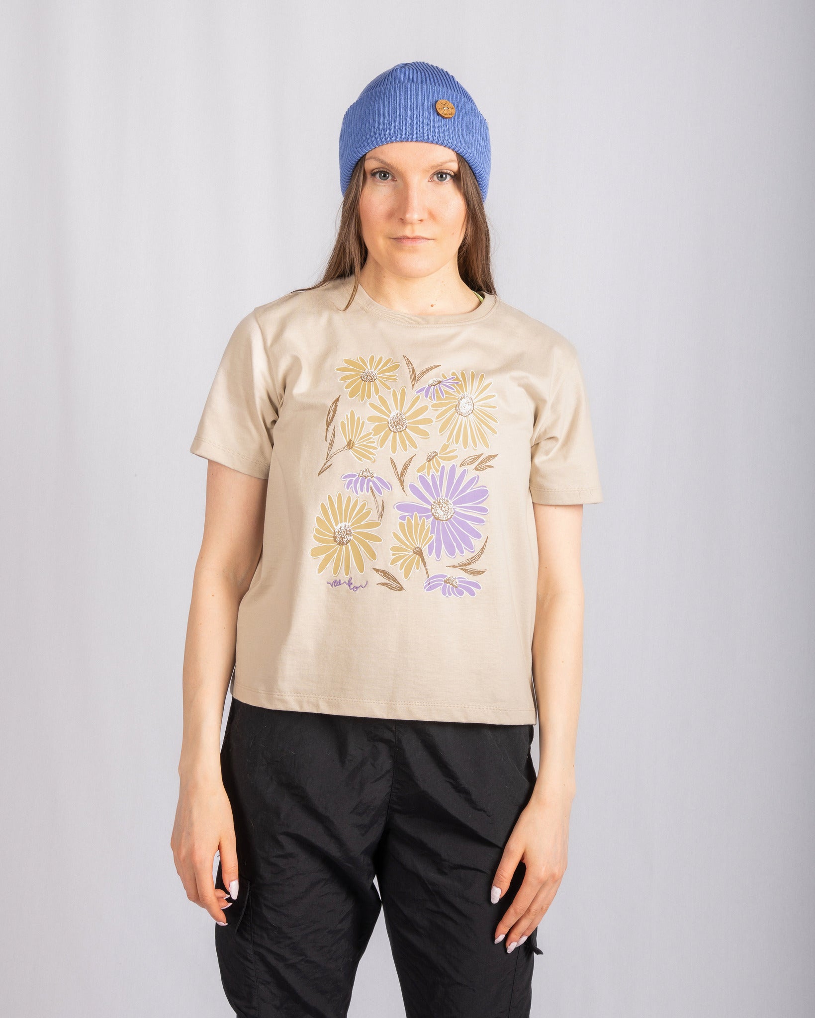 Woman in a blue beanie and floral beige t-shirt stands against a plain light gray background.