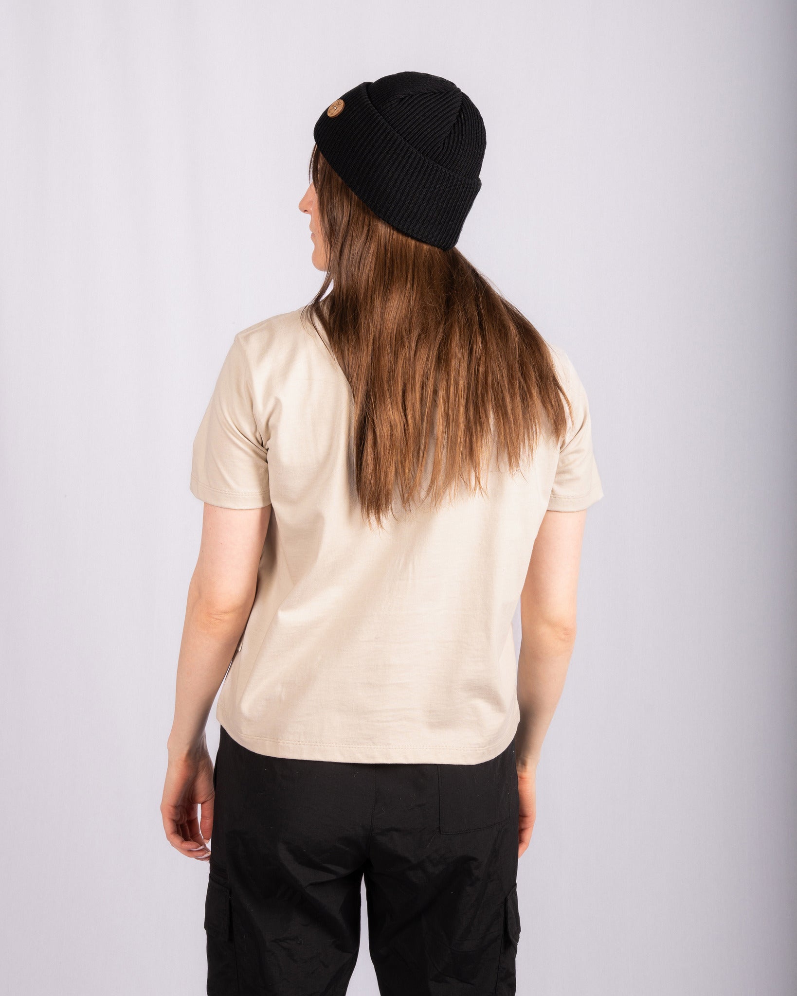 Person facing away, wearing a beige t-shirt, black pants, and a black beanie, against a white background.