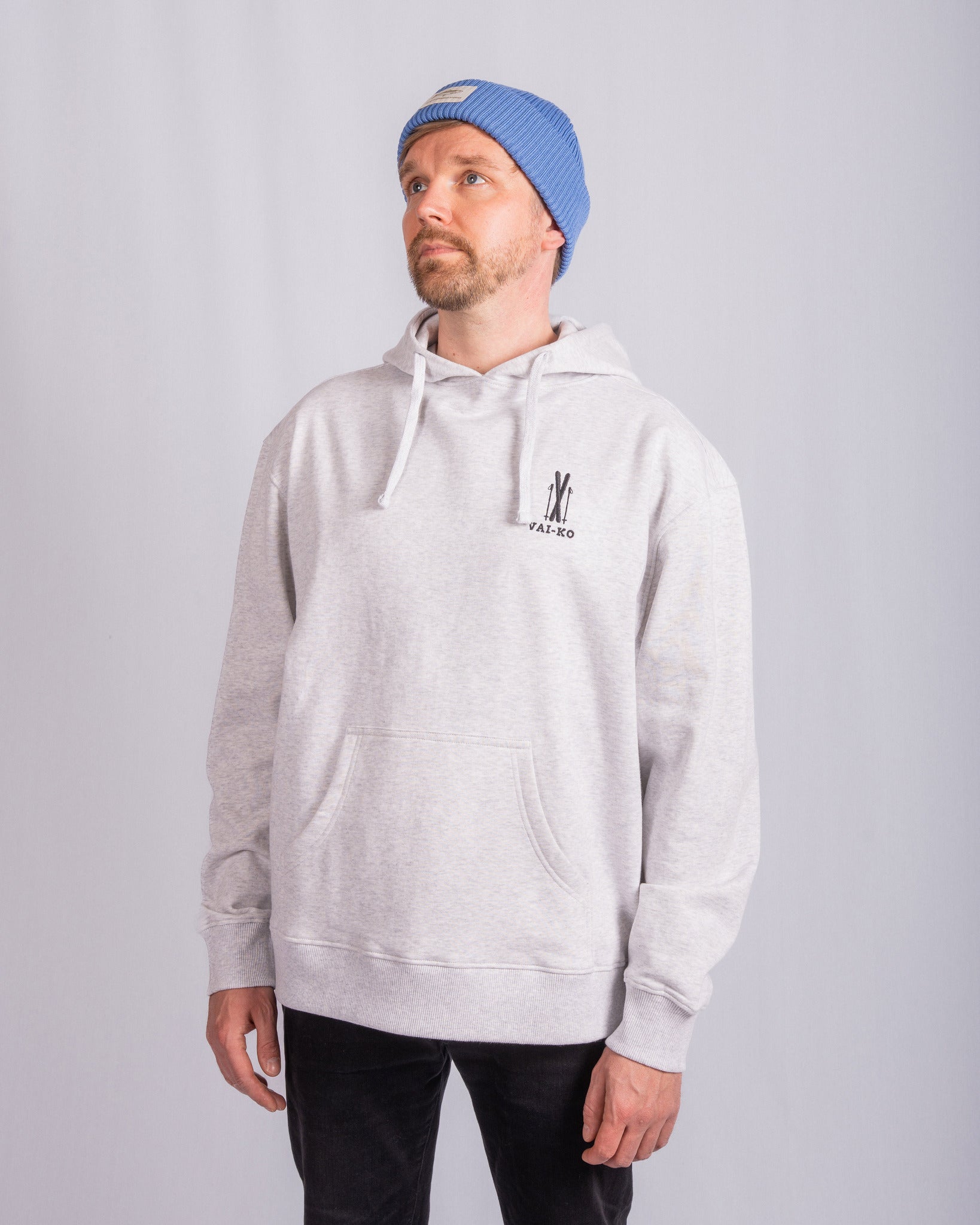 Man in gray hoodie and blue beanie stands against plain light background, looking up and to the side.