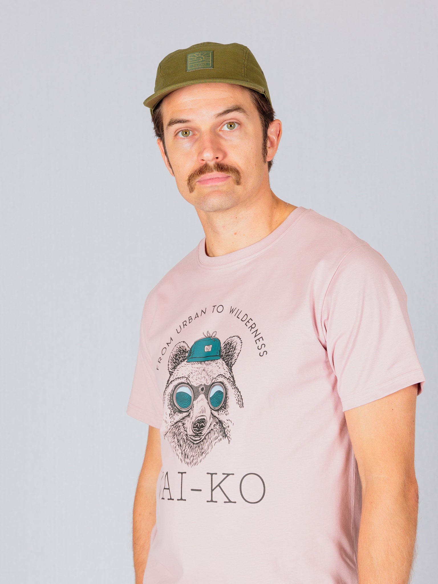 Man with a mustache wearing a green cap and a pink T-shirt with a bear graphic and text.