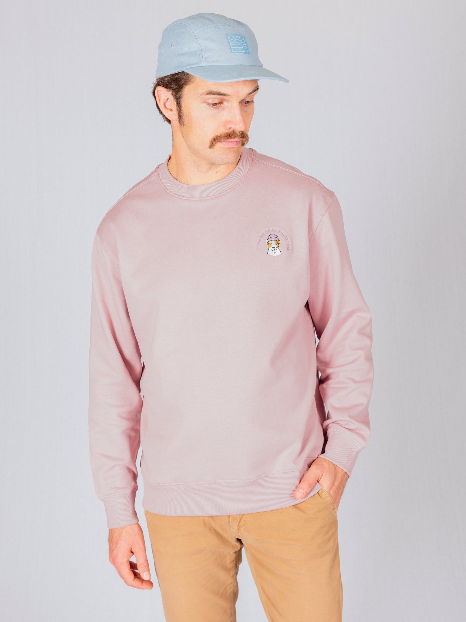 Man in a light pink sweatshirt, tan pants, and a light blue cap stands against a plain light background.