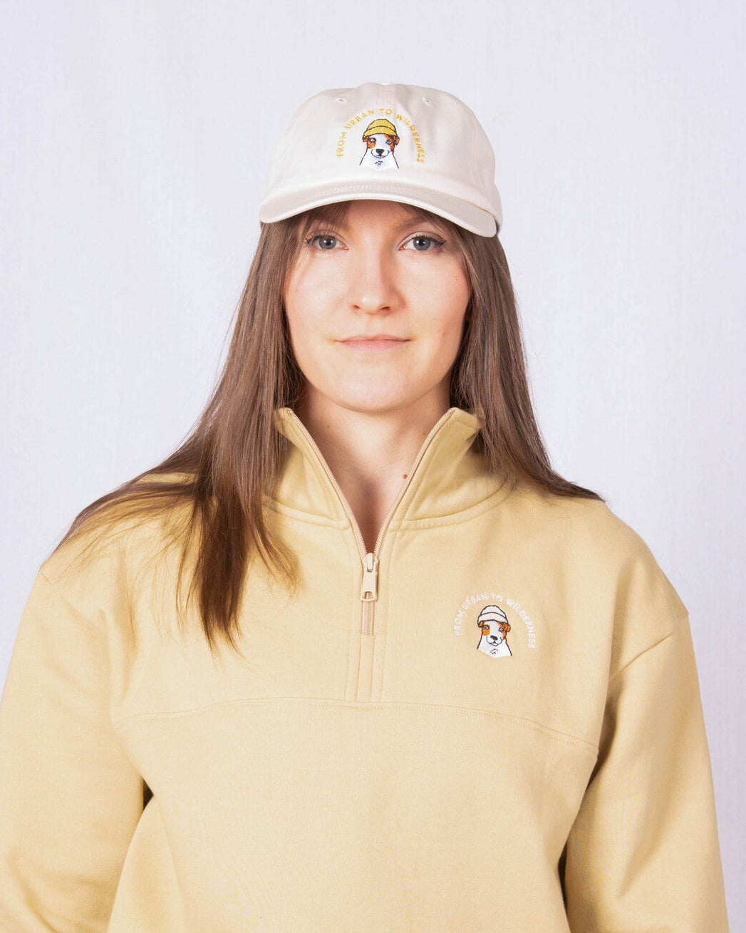 Woman wearing a beige quarter-zip sweatshirt and matching cap, both with a small embroidered logo on the front.