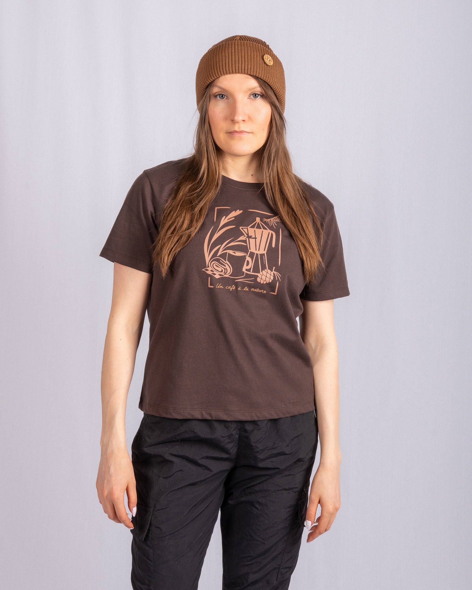 Woman in a brown beanie, brown graphic t-shirt, and black pants standing against a light gray background.