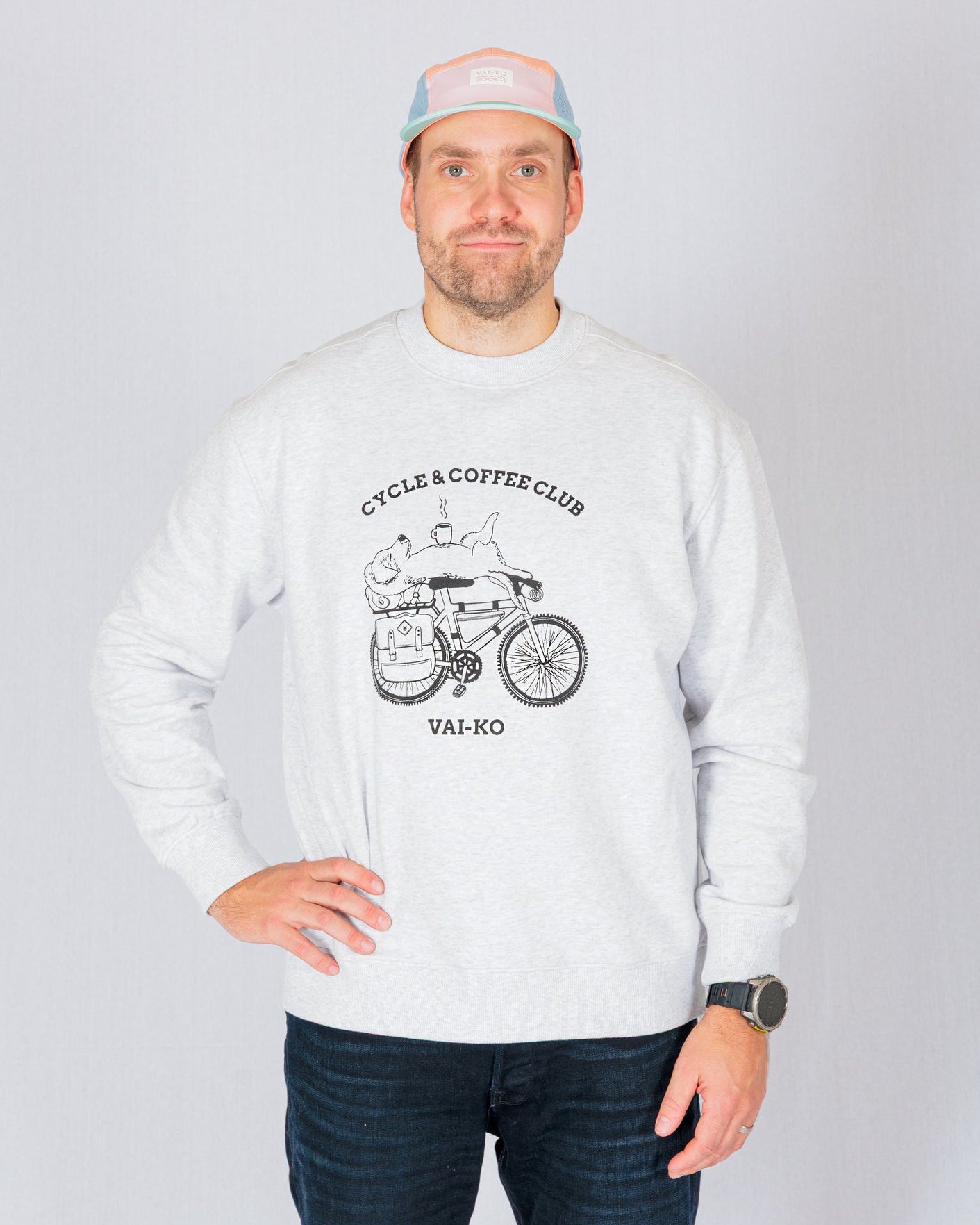 Man in a light sweatshirt with Cycle & Coffee Club graphic, wearing a pastel cap, stands against a gray background.