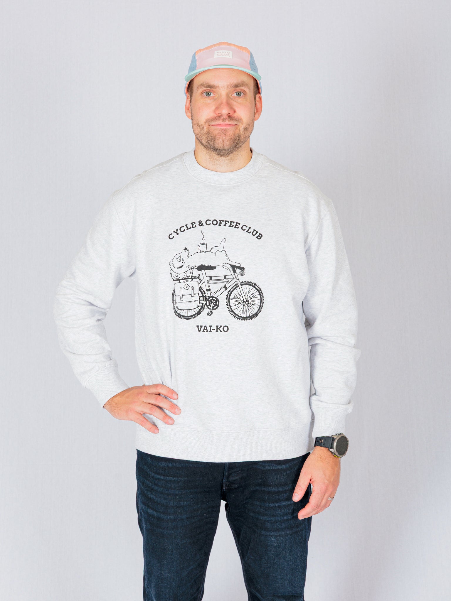 Man in a light sweatshirt with Cycle & Coffee Club graphic, wearing a pastel cap, stands against a gray background.