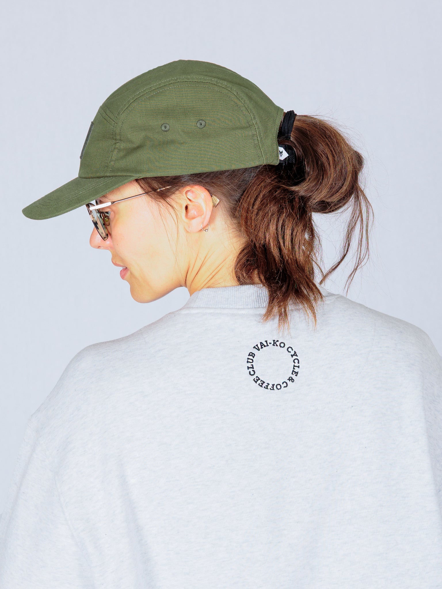 Woman in a green cap and sunglasses, facing away, wearing a light gray shirt with a circular logo on the back.