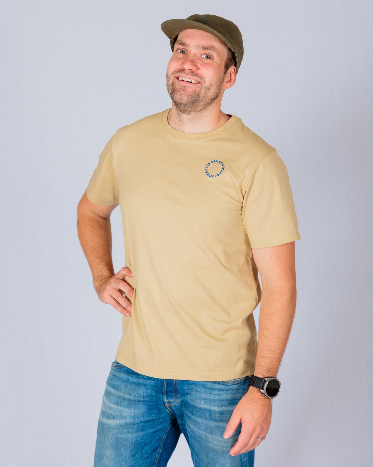 Man in a beige t-shirt, blue jeans, and a green cap smiling with one hand on his hip against a light background.