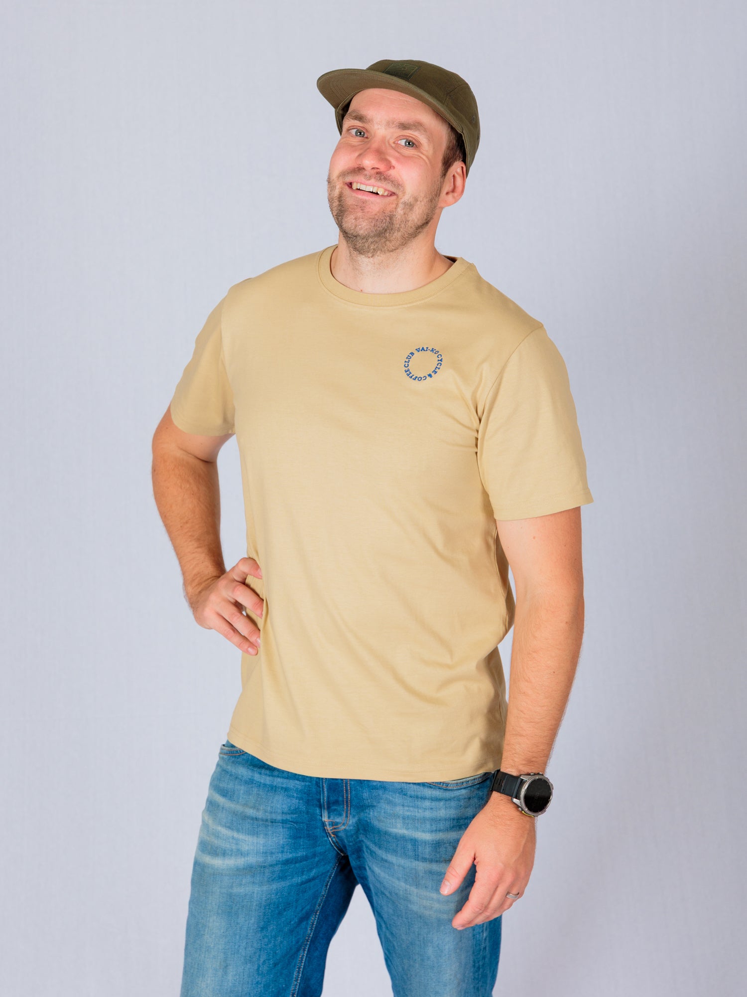 Man in a beige t-shirt, blue jeans, and a green cap smiling with one hand on his hip against a light background.
