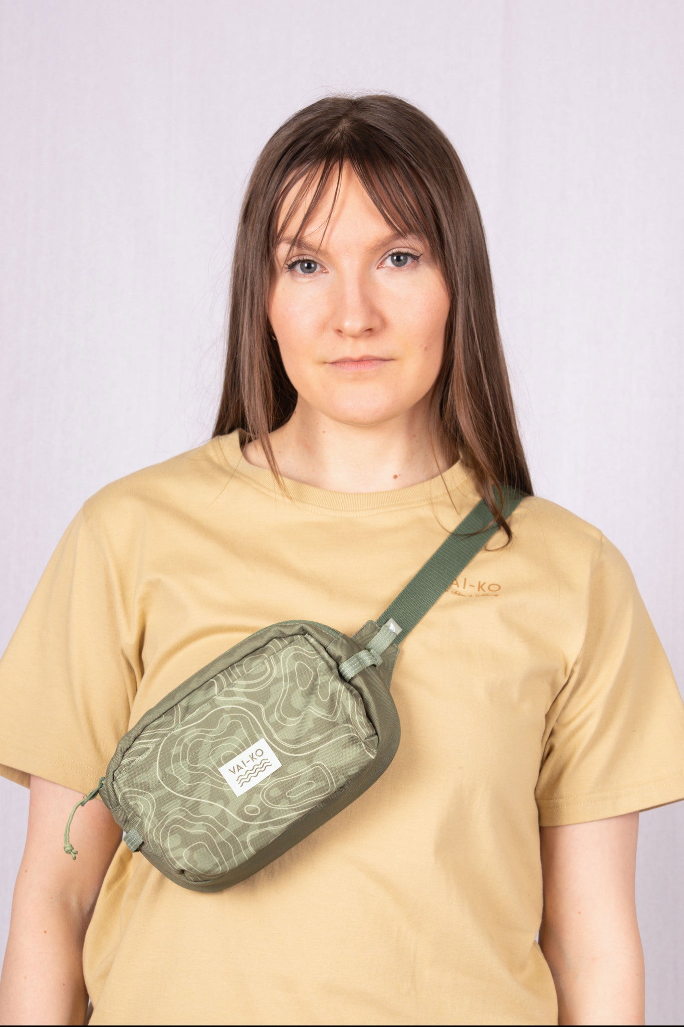 Woman wearing a tan t-shirt and a green crossbody bag, standing against a light background.