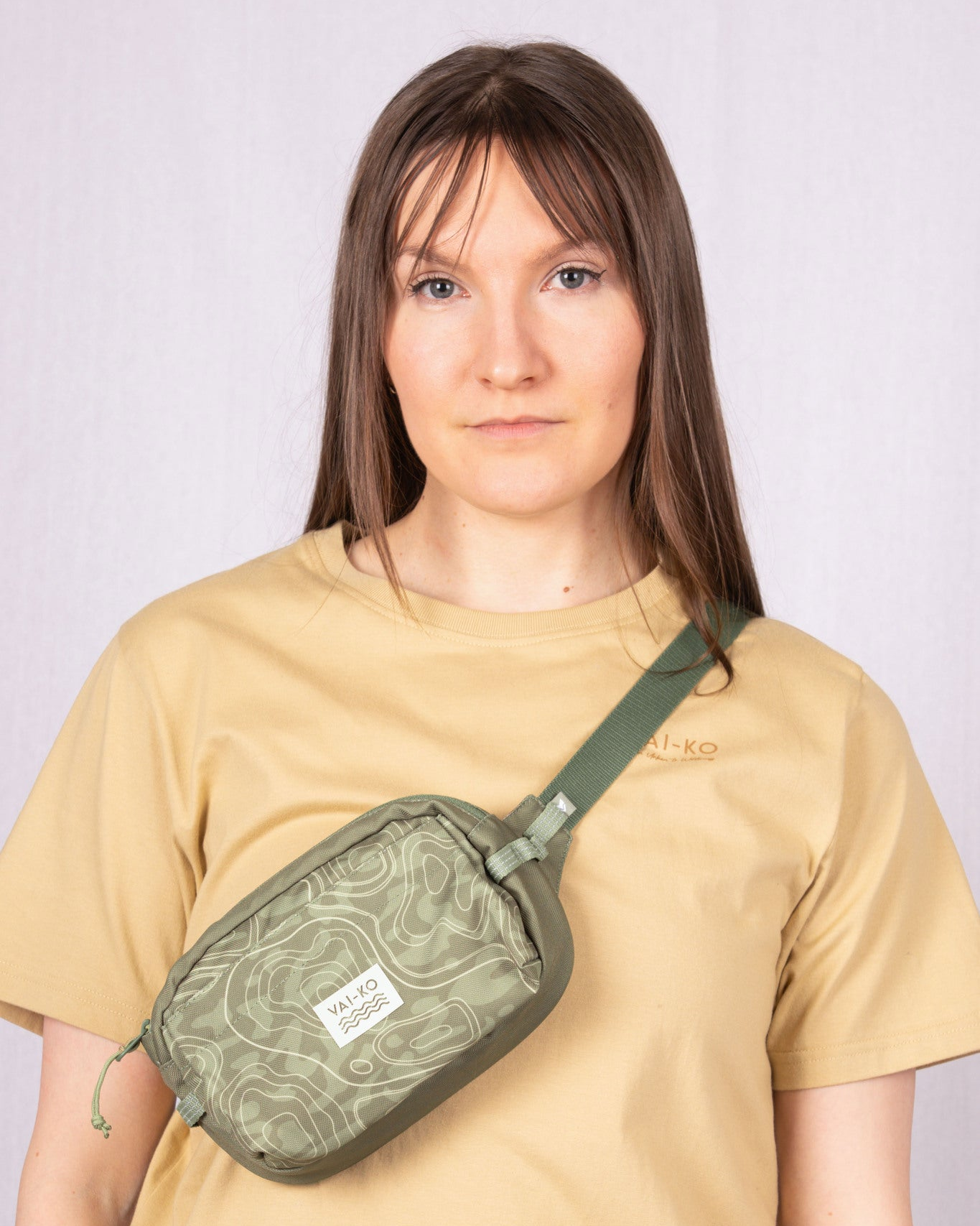 Woman wearing a beige t-shirt and an olive green patterned crossbody bag, standing against a light background.