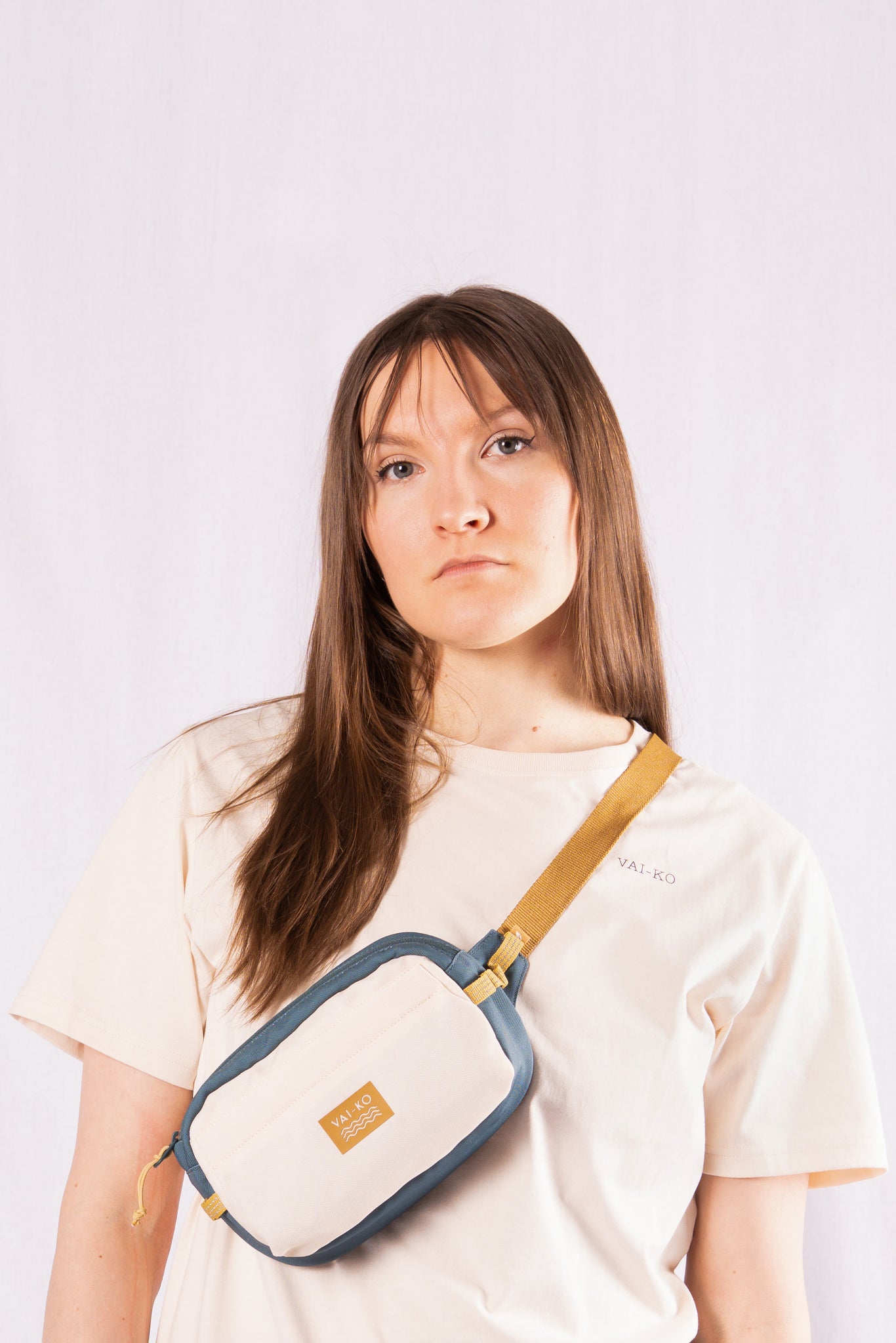 Woman with long brown hair wearing a cream shirt and a crossbody beige and blue bag, standing against a white background.