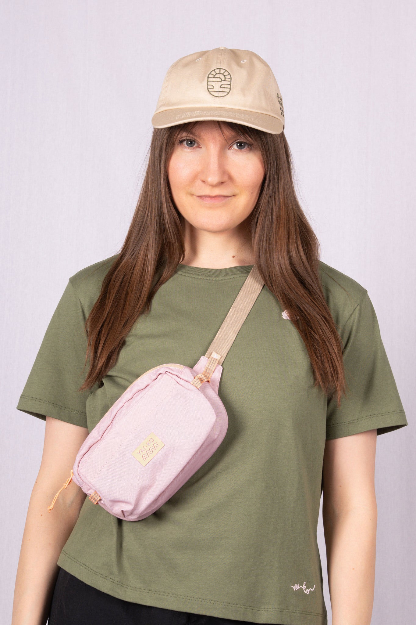Woman in a beige cap, green t-shirt, and crossbody pink bag, standing against a light background.