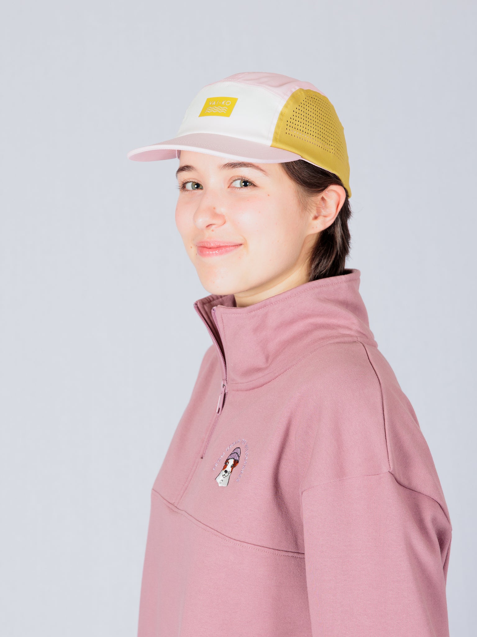 Person in a pink pullover and pastel baseball cap, smiling and standing against a light gray background.