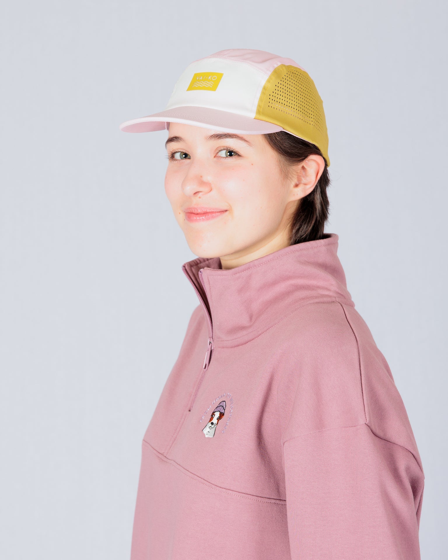 Person in a pink pullover and pastel baseball cap, smiling and standing against a light gray background.