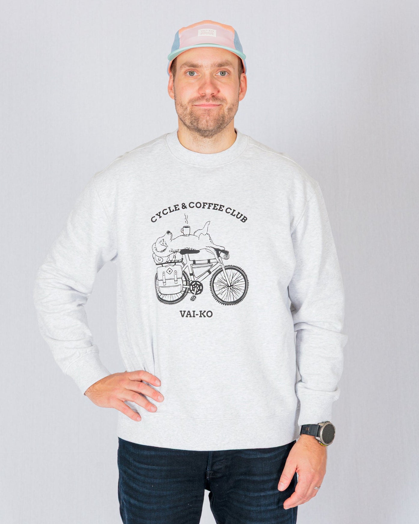 Man in a light sweatshirt with “Cycle & Coffee Club” graphic, wearing a pastel cap, stands against a plain background.