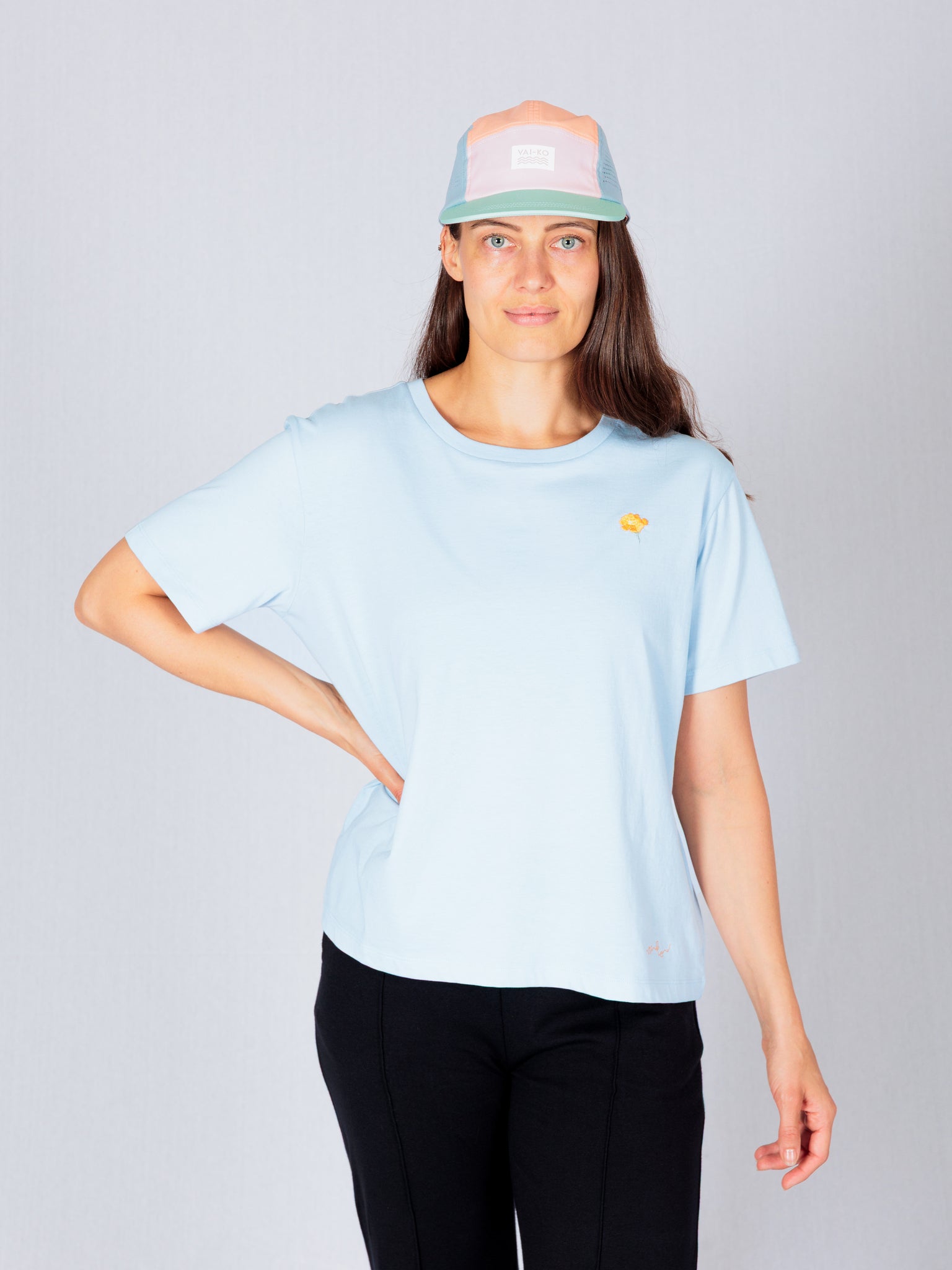 Woman in a light blue t-shirt and pastel cap stands with one hand on her hip against a plain background.