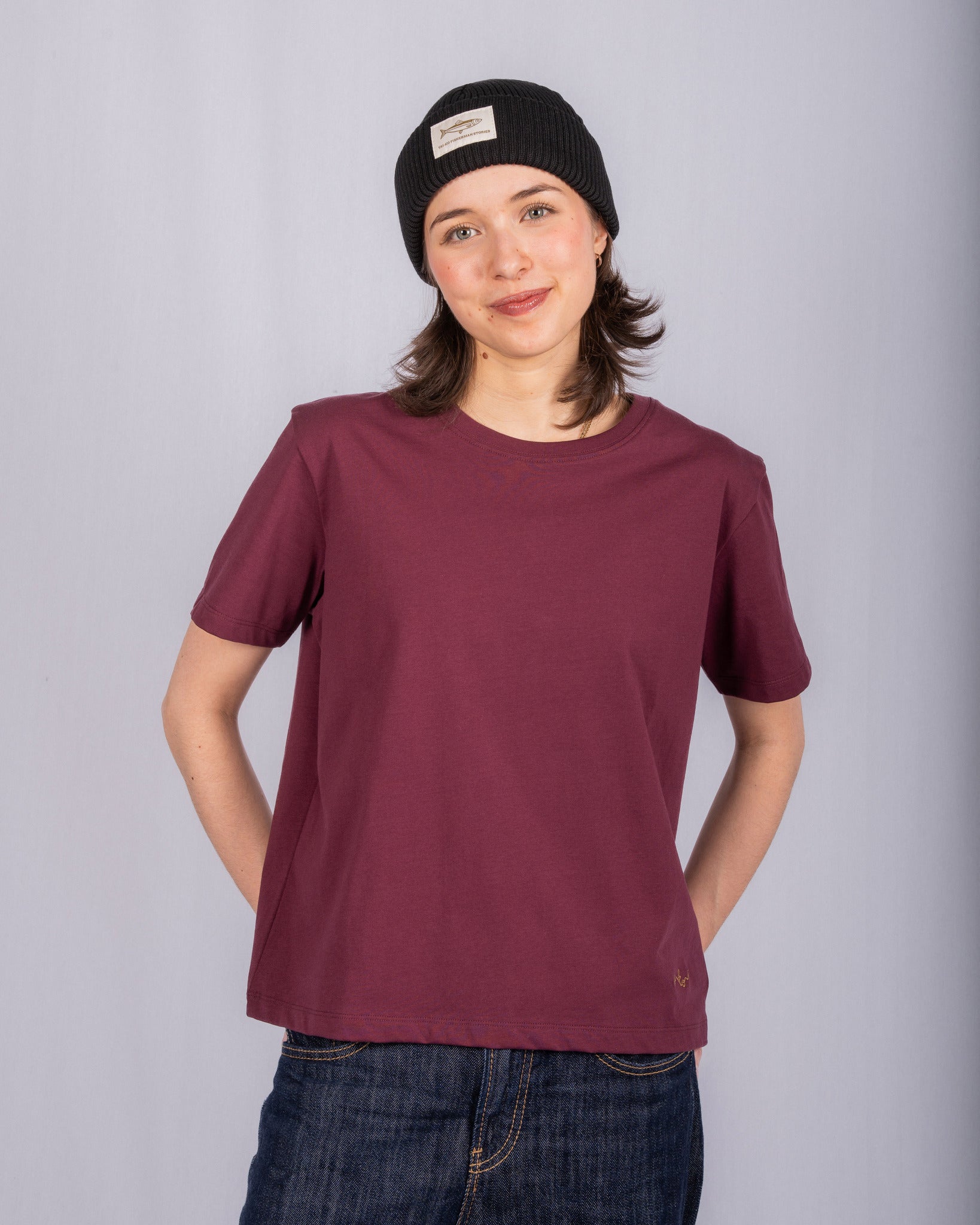 Person in a maroon t-shirt, jeans, and black beanie stands smiling against a plain light gray background.